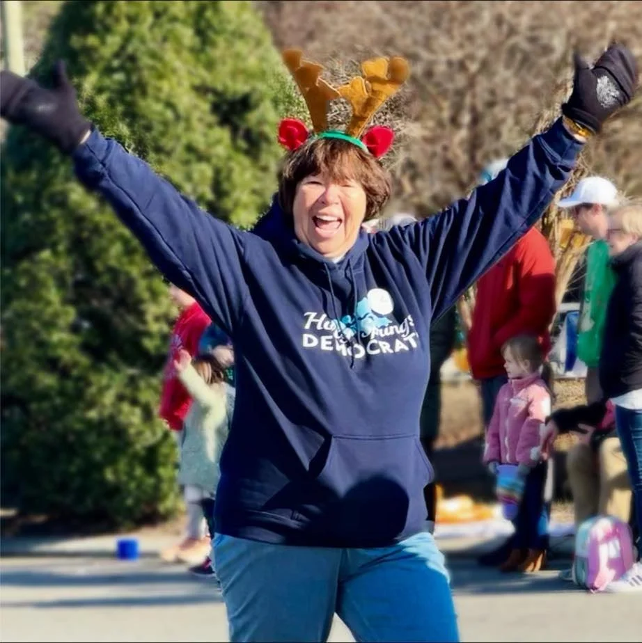 An older woman smiles and cheers as she participates in a parade. She is wearing a navy blue sweatshirt, an festive headband with antlers, and is throwing her arms in the air.