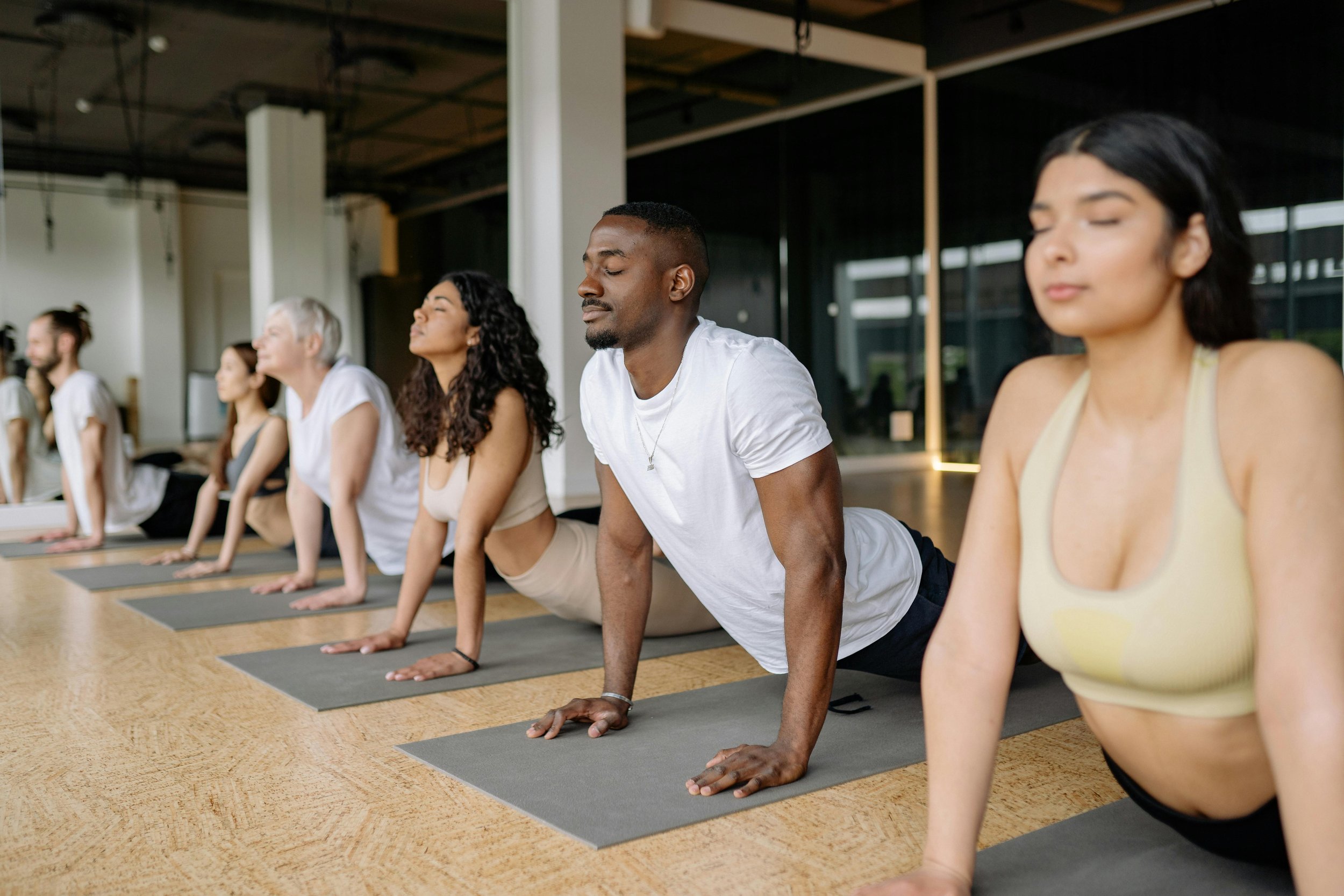 Group of diverse people practicing yoga in a studio, performing upward-facing dog pose on mats in a well-lit room with large windows.