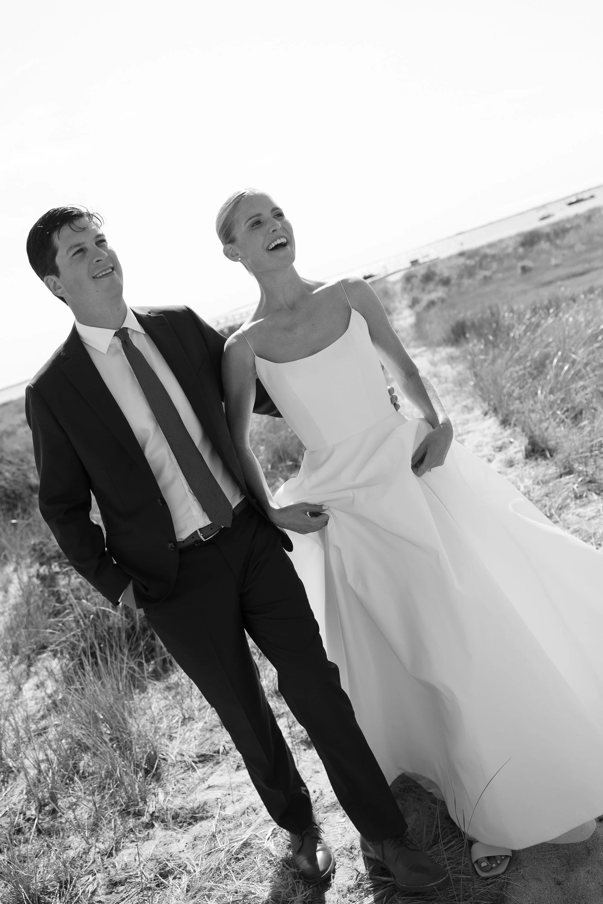 Wedding invitation surrounded by white and cream roses, green leaves, and wedding rings in black rings boxes on a white surface.