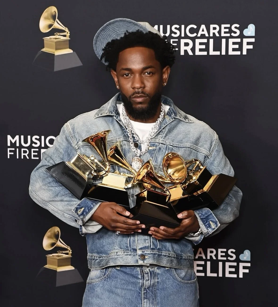 Male musician holding multiple gold Grammy awards at Musicares Relief event, wearing denim jacket and jewelry.
