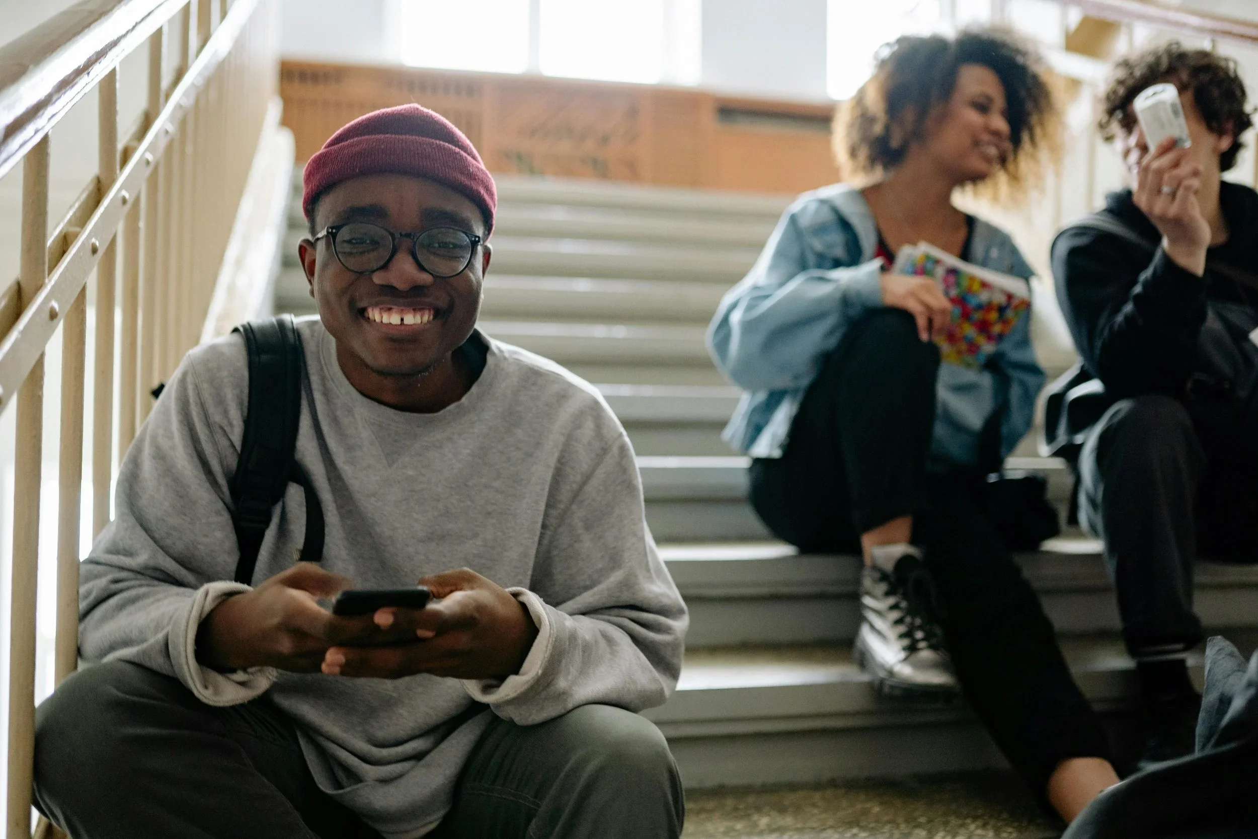 Three young adults sitting on indoor stairs, smiling and engaging with their phones, in a well-lit, casual environment.