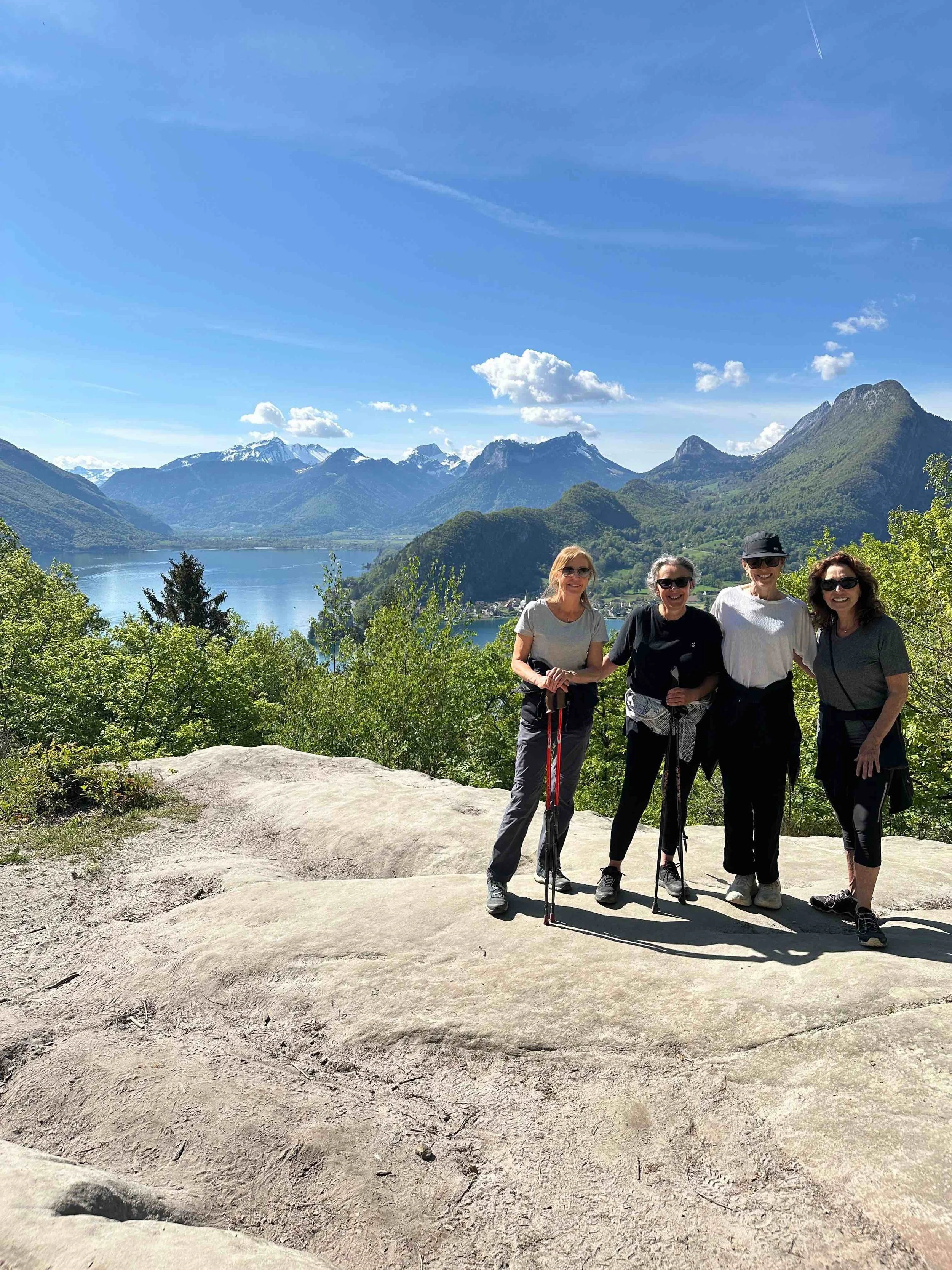Four women standing on a rocky surface with a mountain lake and snow-capped mountains in the background, under a bright blue sky with a few clouds.