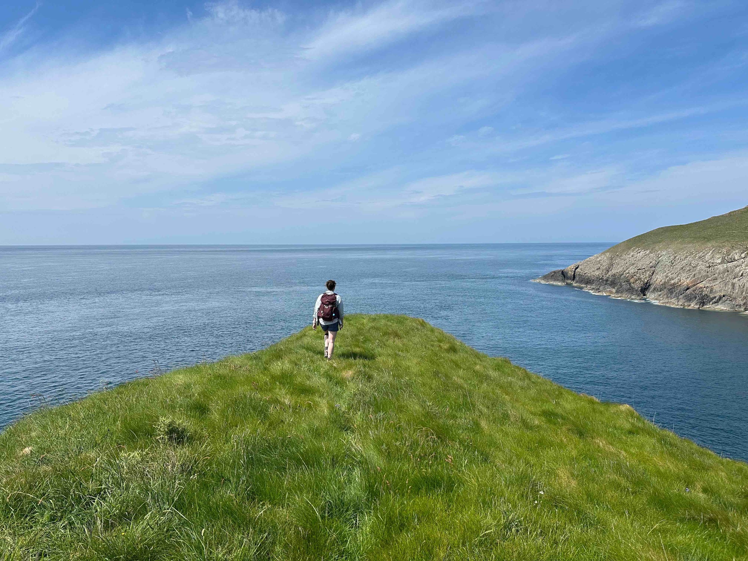A person with a backpack walking along a grassy path on a cliffside overlooking the ocean, with blue sky and clouds above.