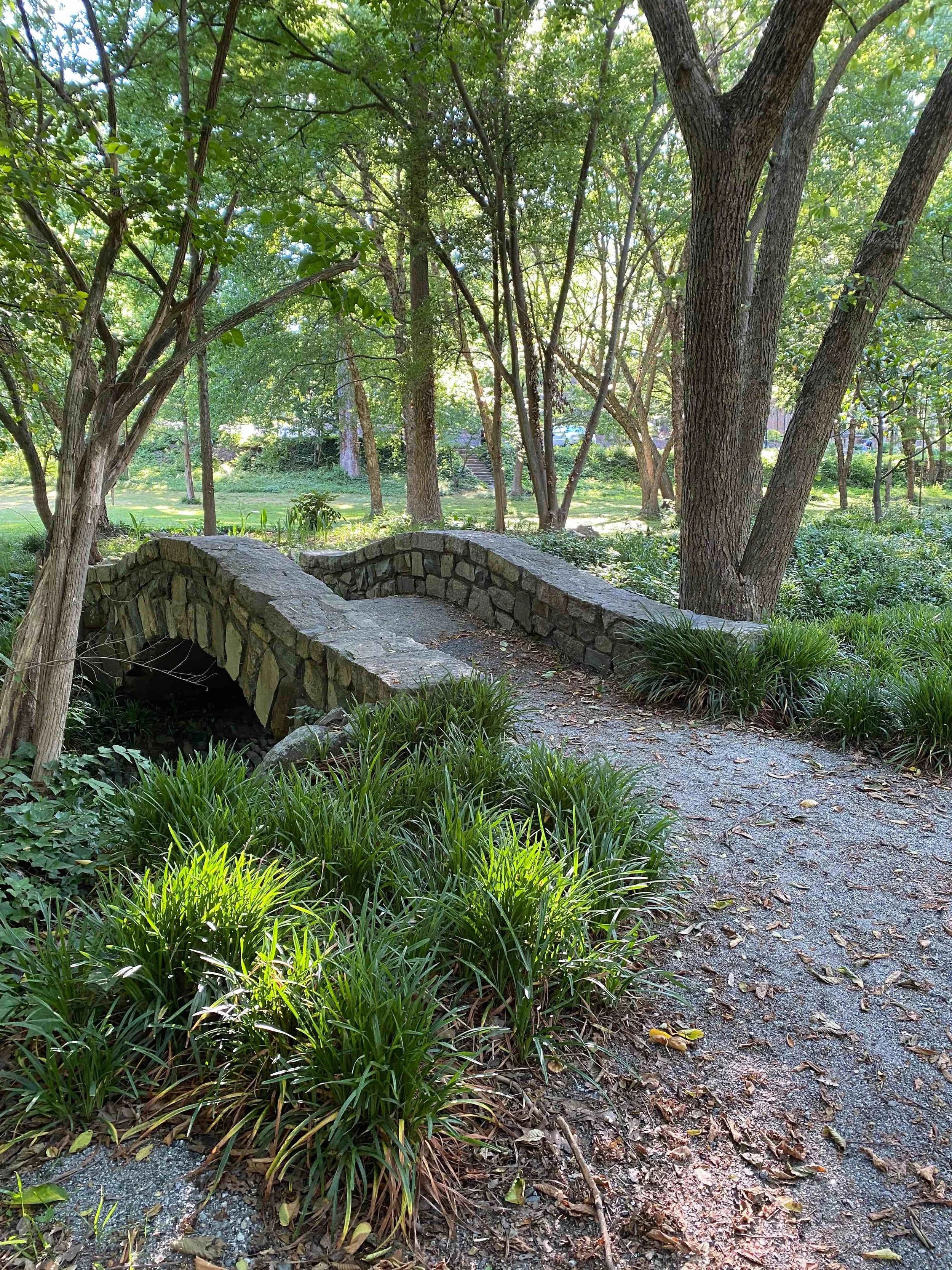 A stone footbridge in a lush green park with tall trees, bushes, and a gravel pathway