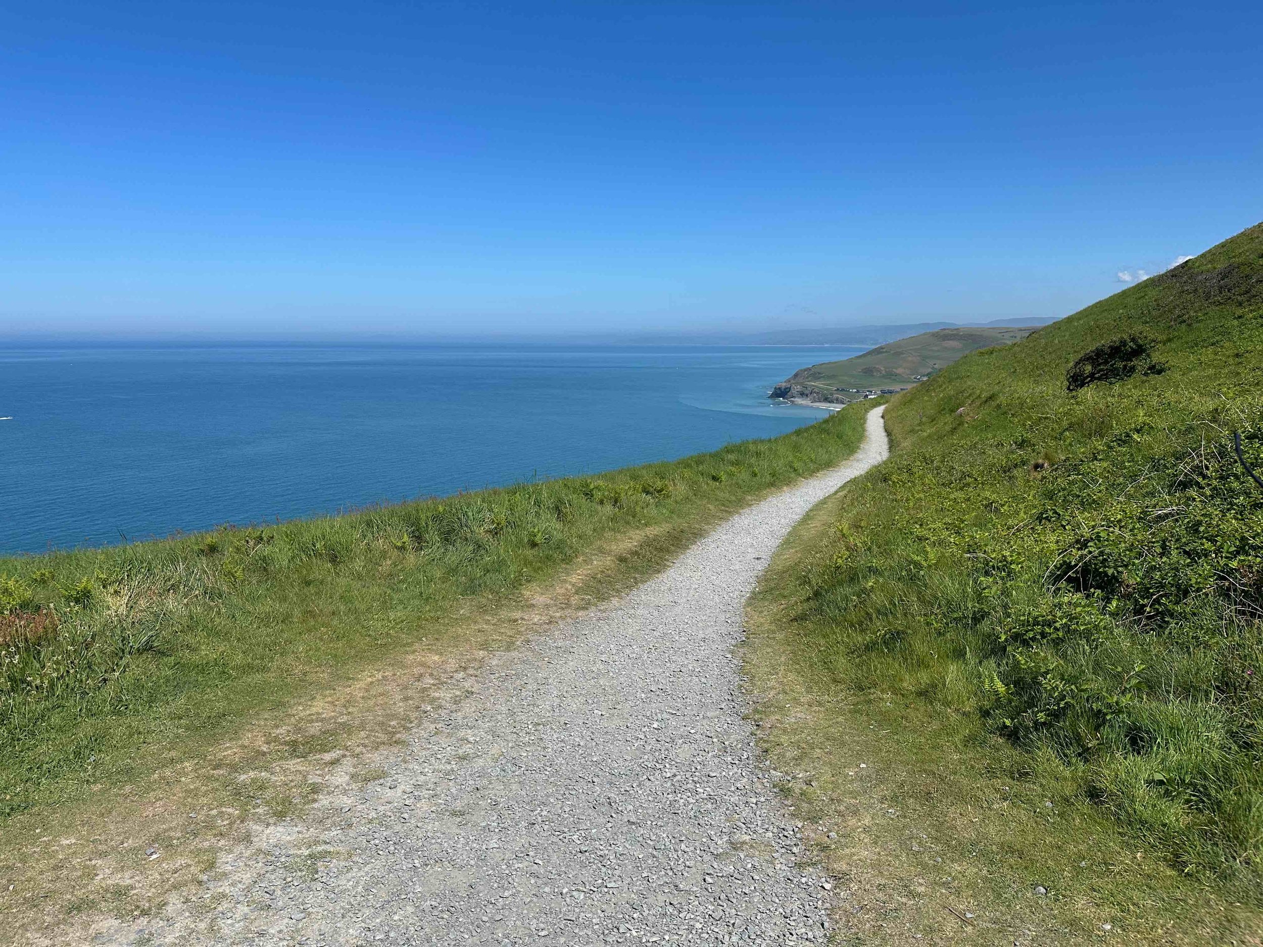 A gravel path in-between green grass and a hill winds next to a blue sea.