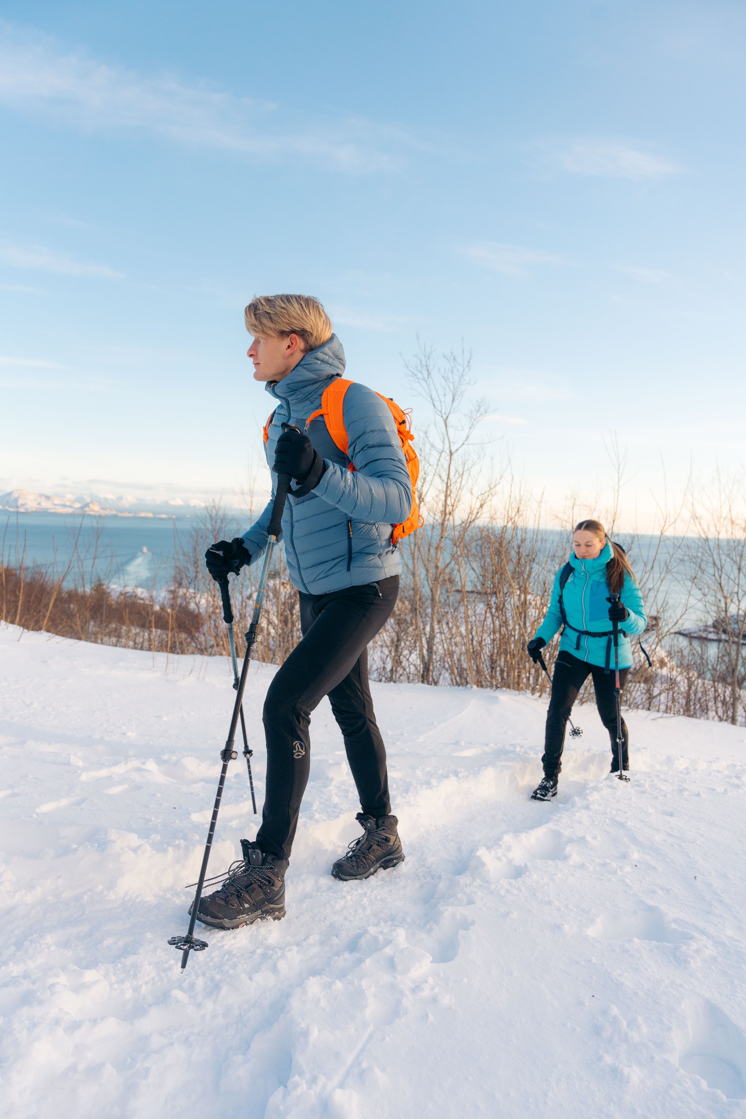 Personas caminando con equipo de senderismo en la nieve, con bastones de trekking y mochilas, cerca de un paisaje nevado con árboles y un fondo de mar. Cielo despejado.