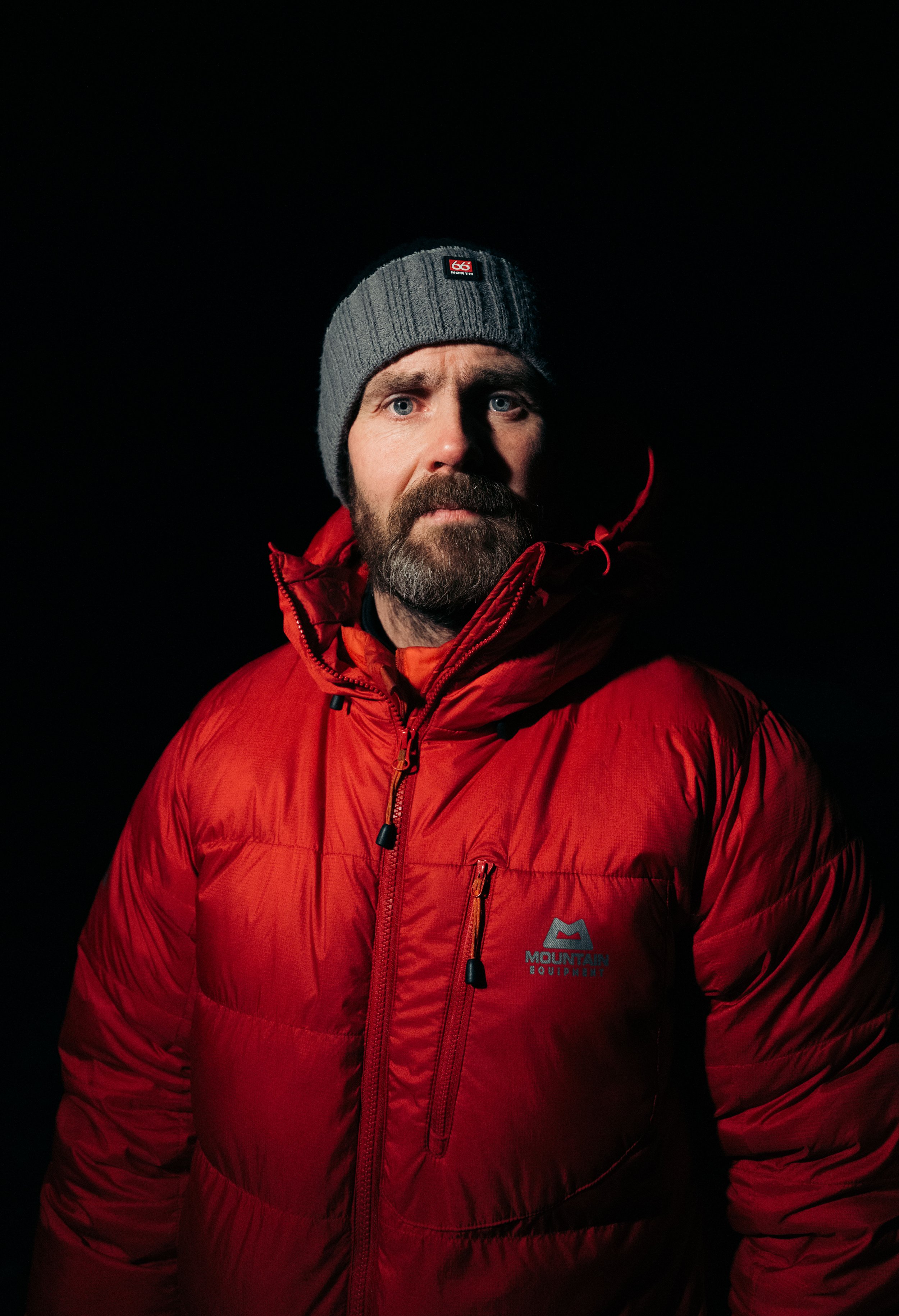 Hombre con chaqueta roja y gorro gris sobre fondo oscuro.