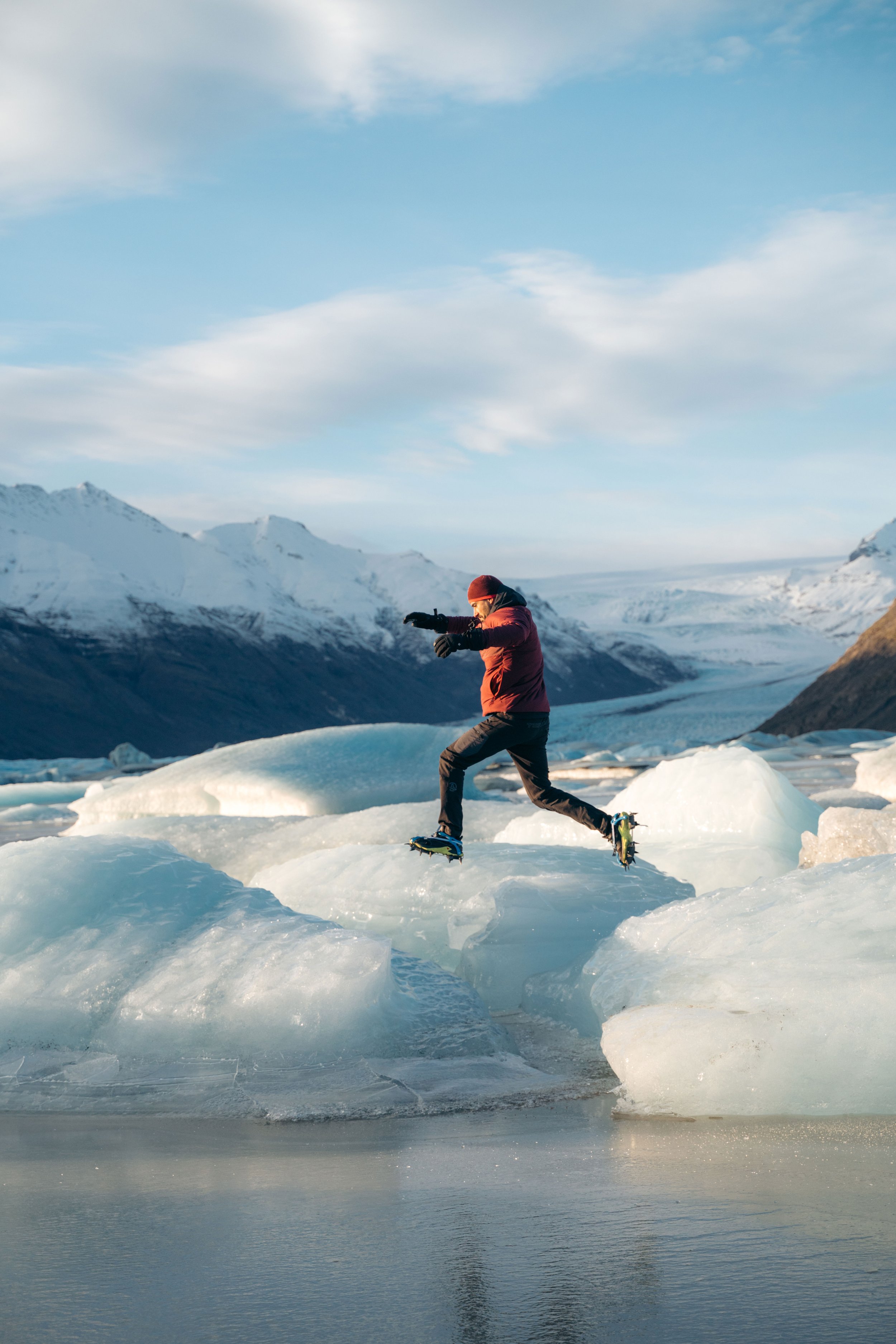 Persona saltando sobre bloques de hielo en un paisaje nevado, con montañas al fondo.