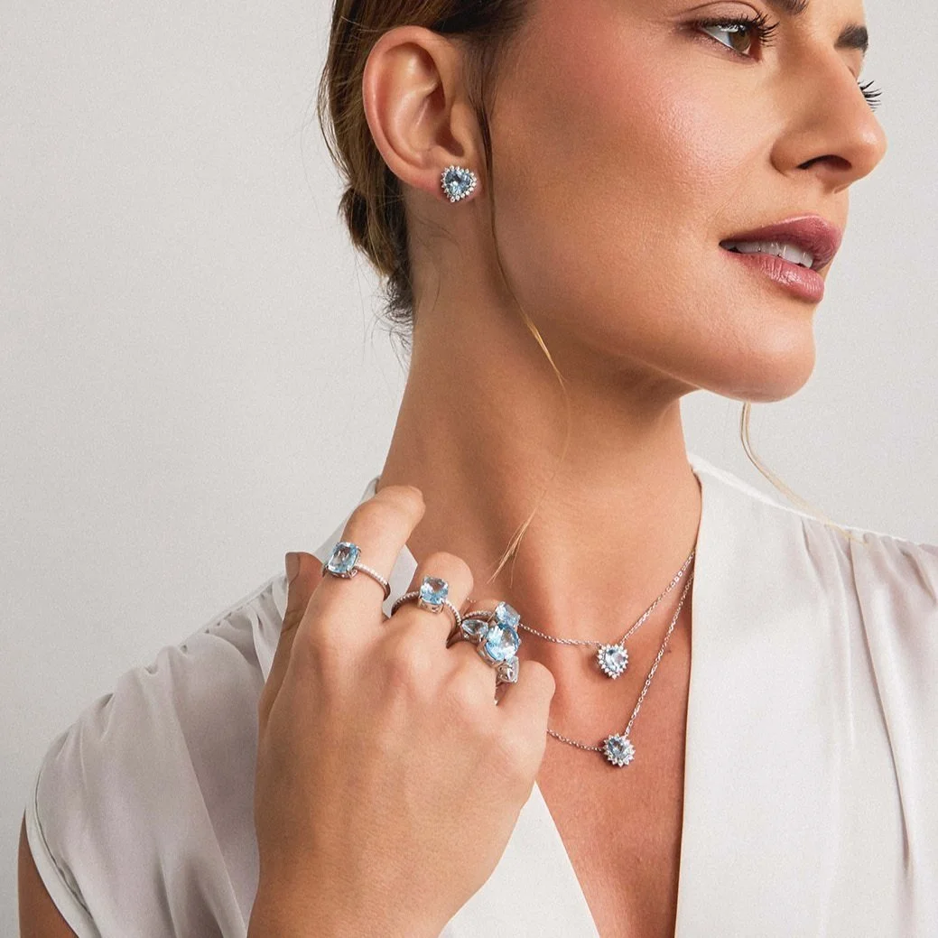 Mulher vestida com roupas brancas, usando brincos, colares e anéis com pedras preciosas azuis e brancas, posando contra fundo branco.