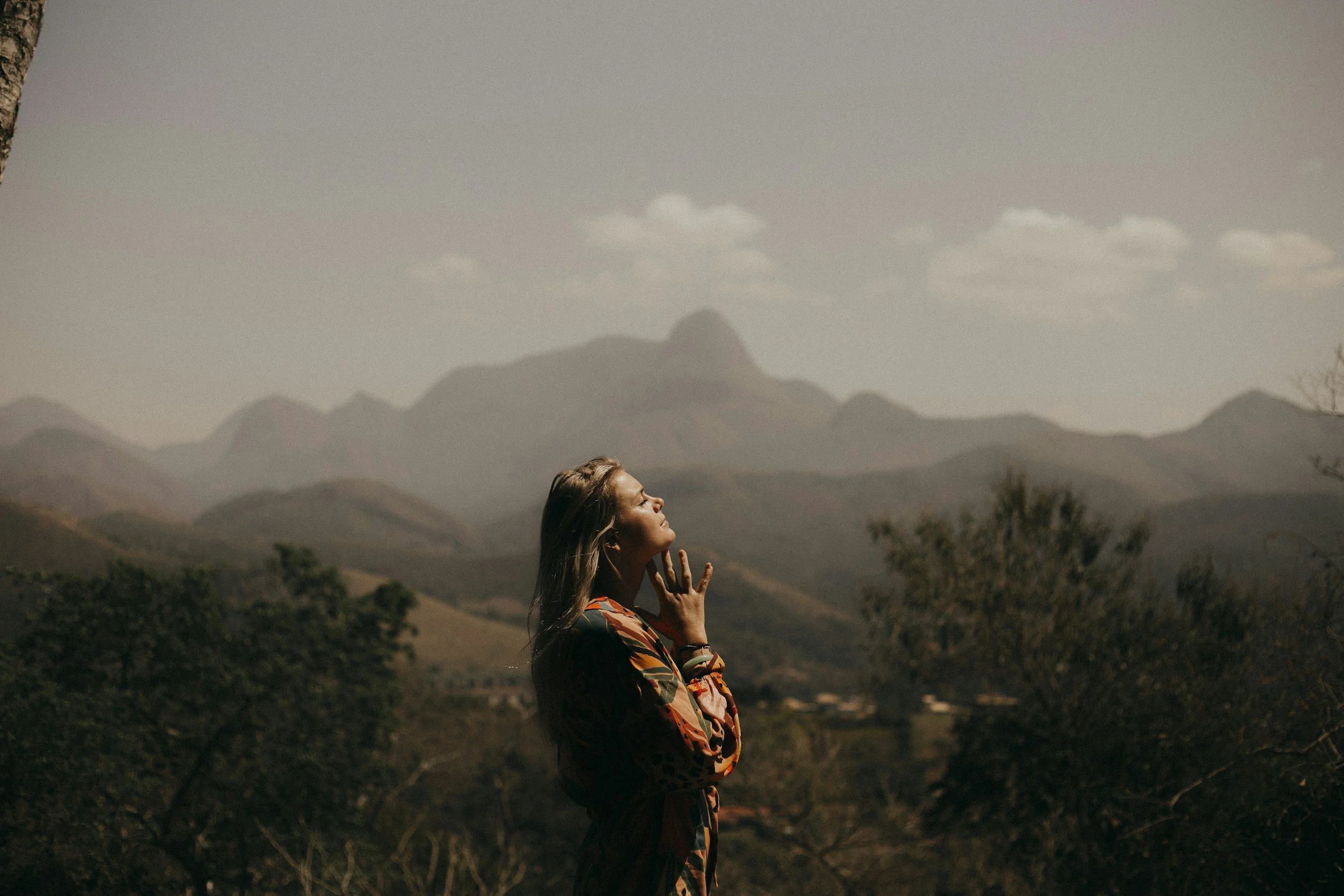Woman in colorful dress standing outdoors, eyes closed, with mountains in background