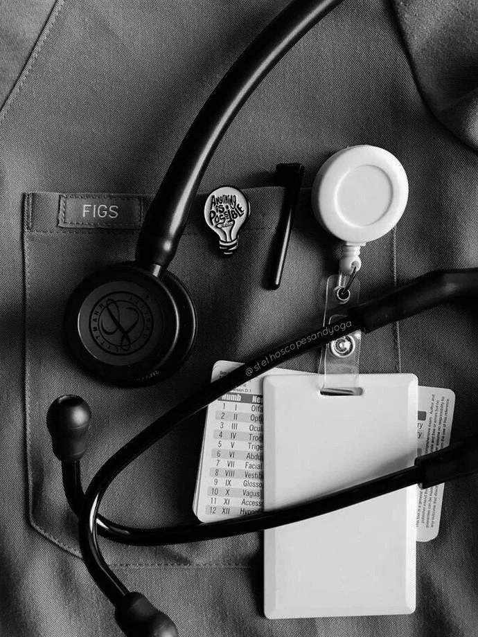Close-up of a medical professional's outfit with a stethoscope, ID badge, and light bulb pin. The name "FIGS" is visible on the pocket.