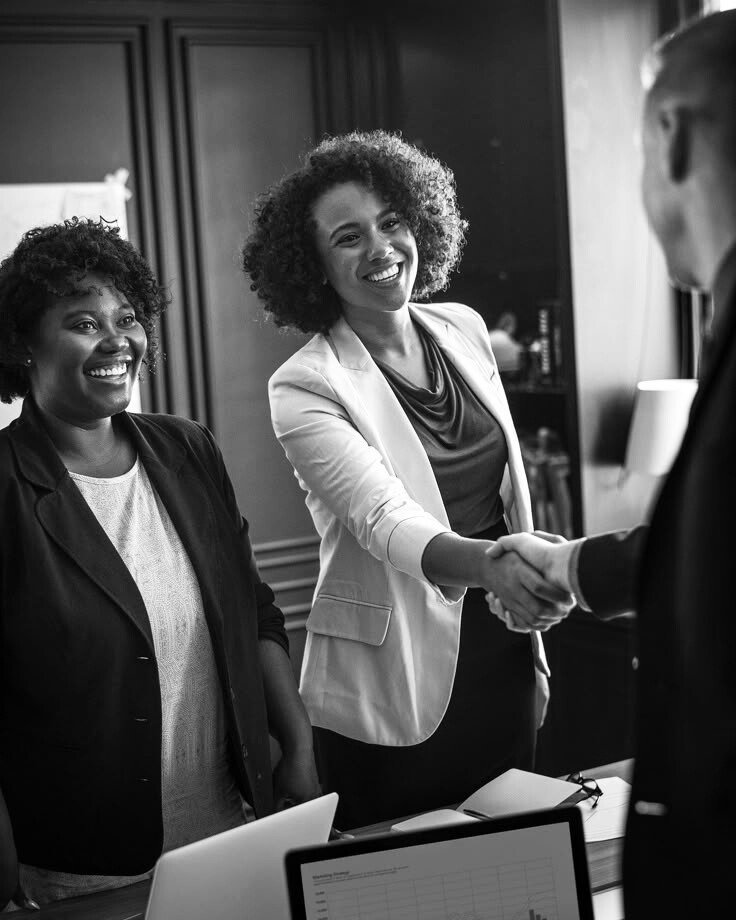 Two women in business attire smiling and shaking hands with a man in an office.