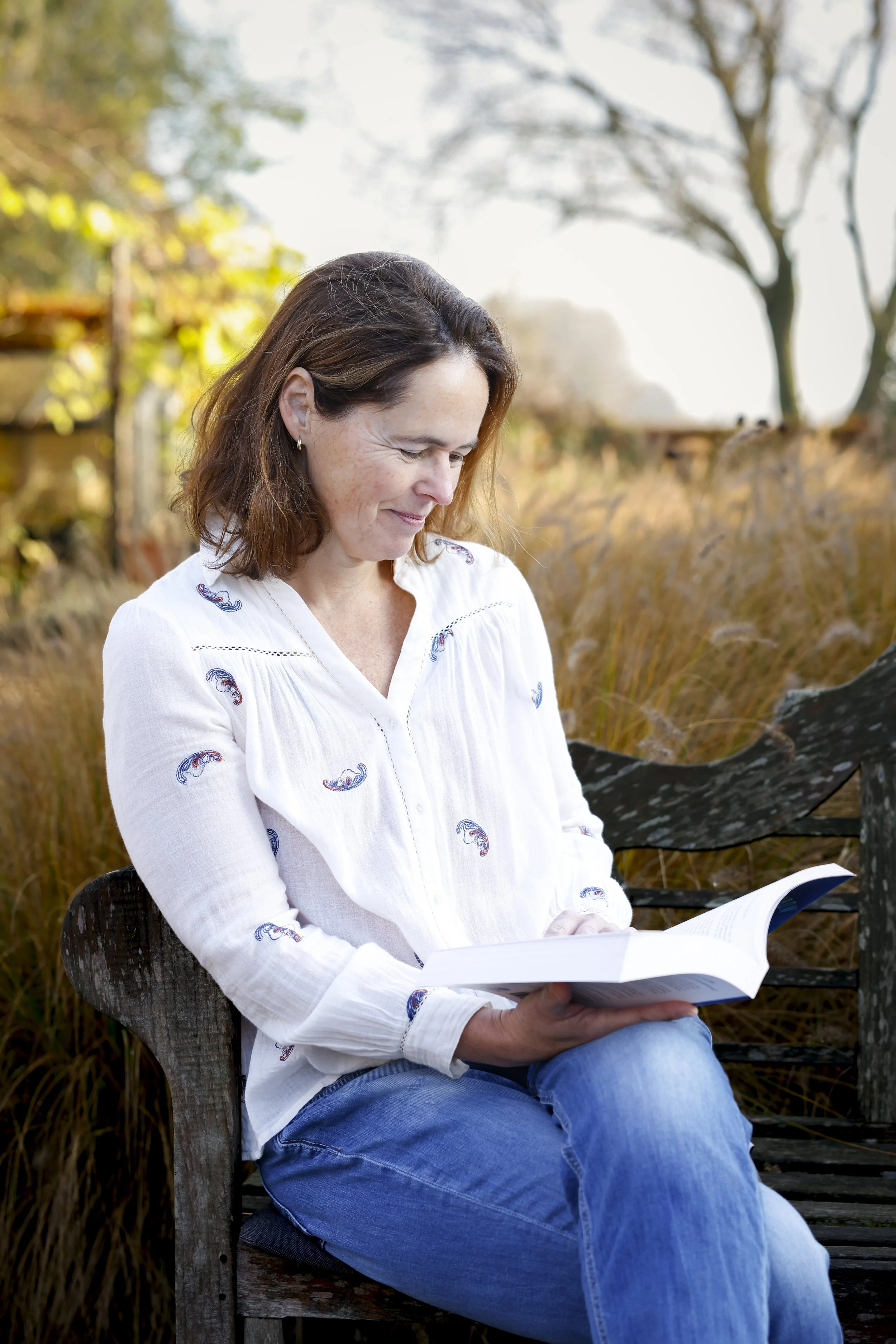 Vrouw zit buiten op een houten bank en leest een boek, met hoge grassen en bomen.