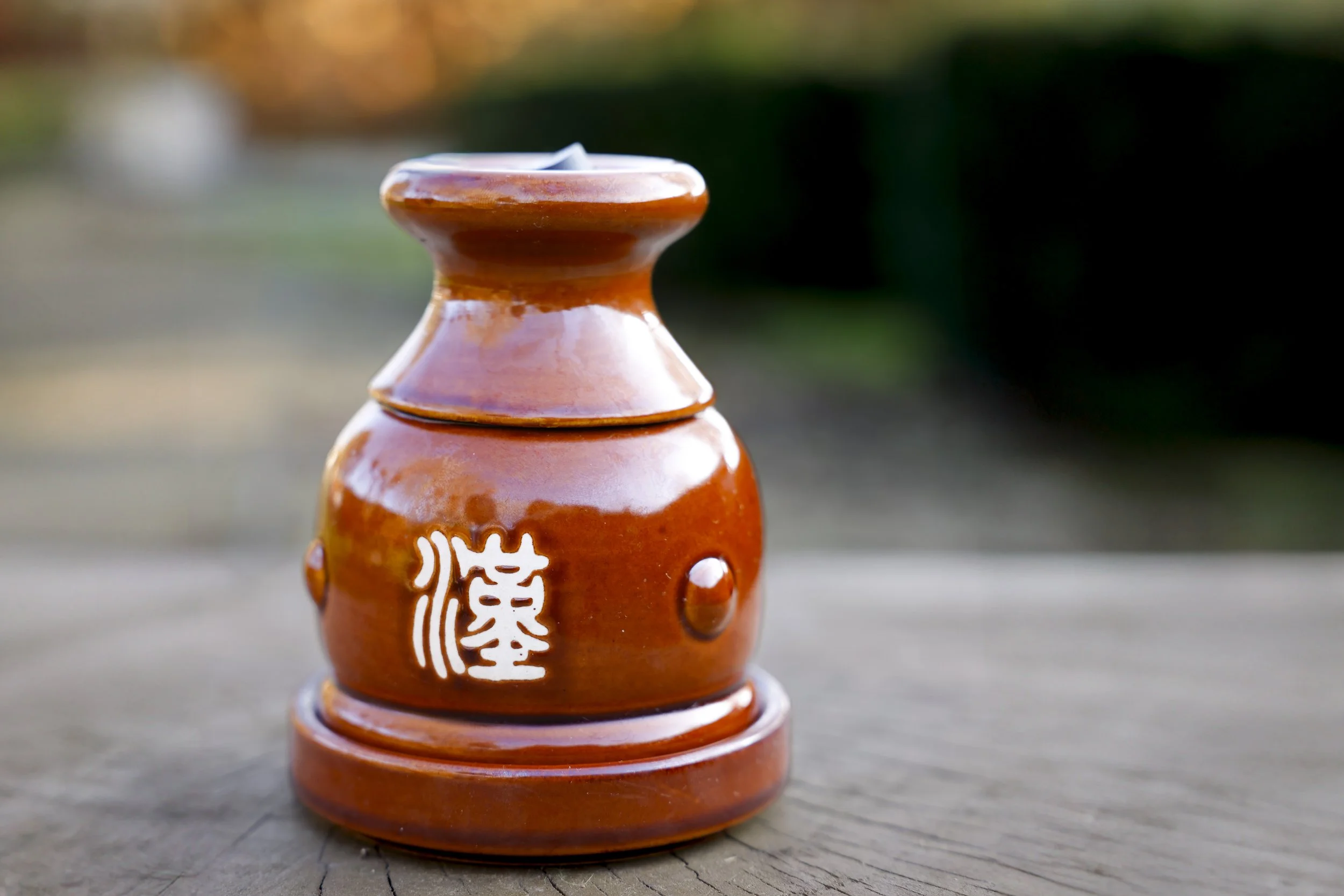 Brown ceramic Chinese incense holder with white Chinese characters, placed on a wooden surface outdoors with a blurred natural background.