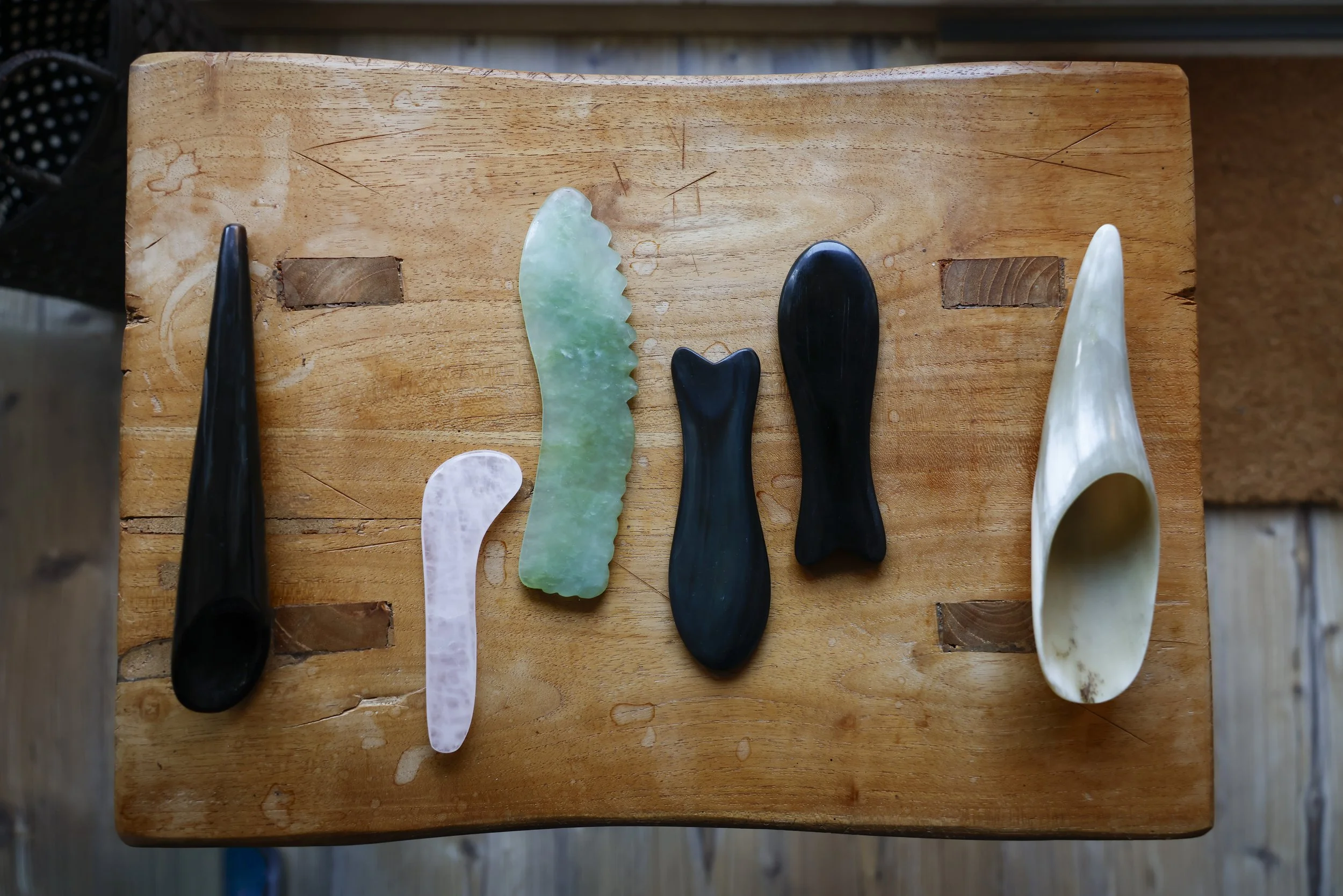 Six carved jade and stone utensils, including a white spoon, a white curved scraper, a green jade knife, a small black fish-shaped piece, a black eggplant-shaped piece, and a white shell-shaped spoon, are arranged on a wooden table.