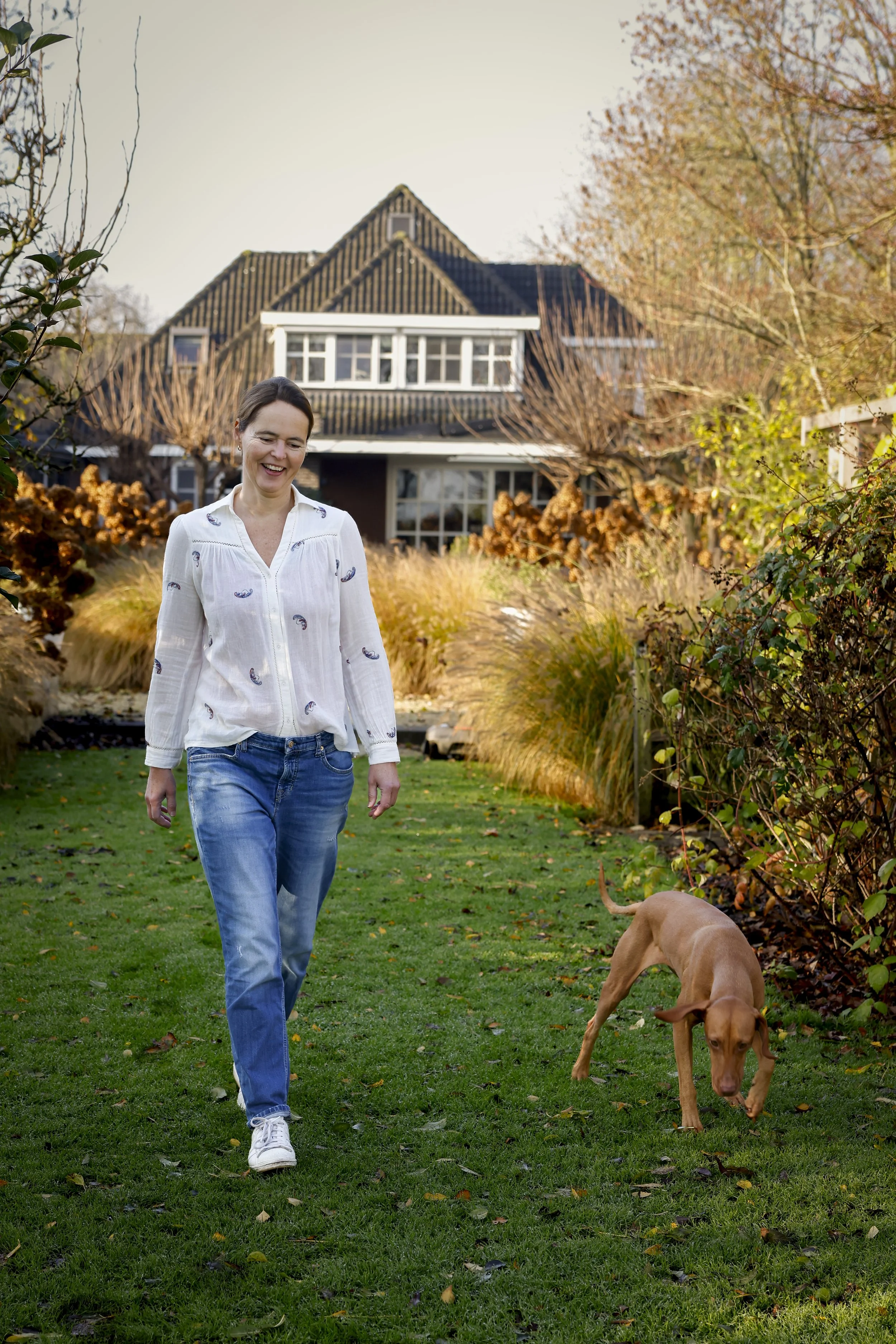 Vrouw loopt over groen gazon met bruine hond voor huis met herfstbomen.