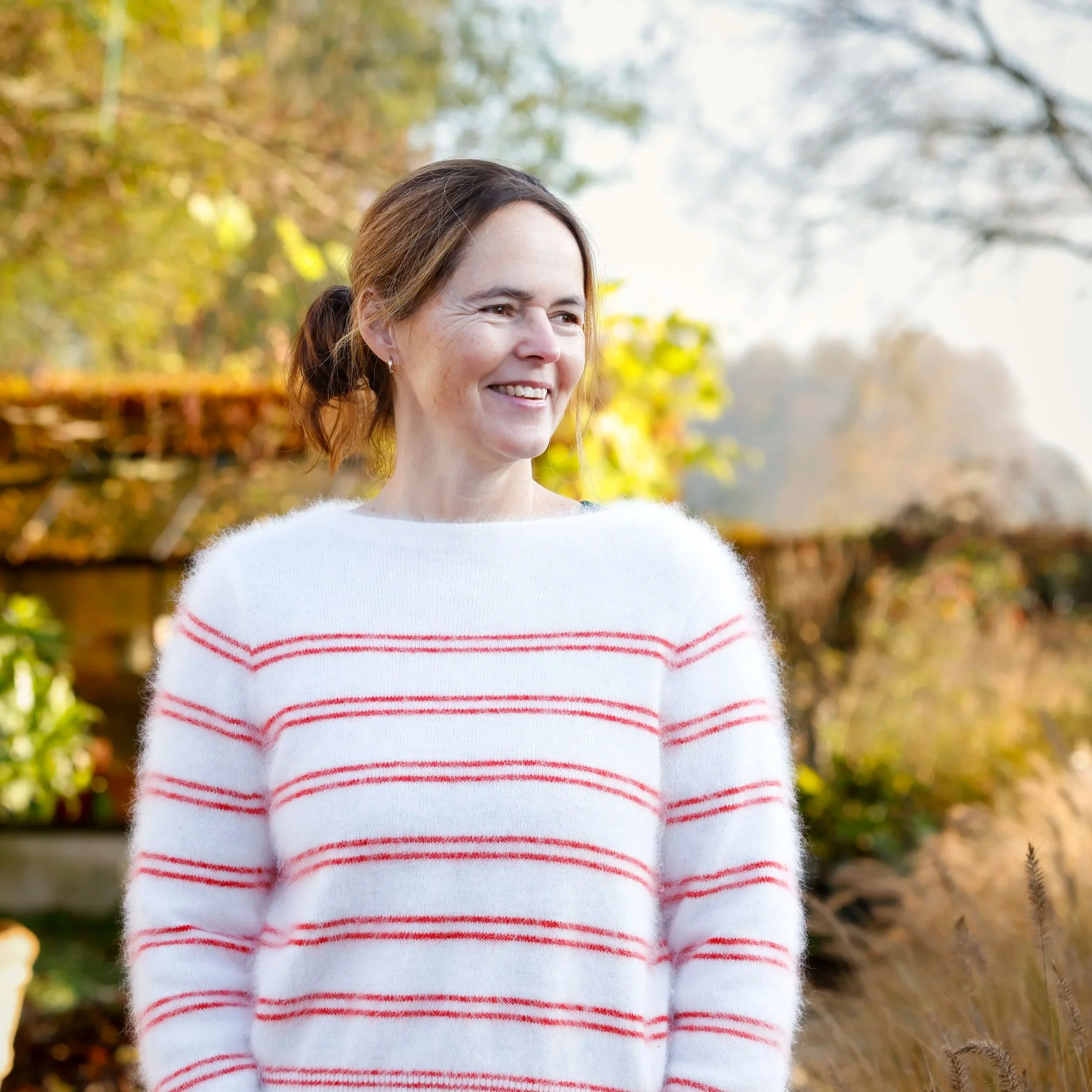 Woman smiling outdoors in autumn, wearing a white sweater with red stripes, with trees and fall foliage in the background.