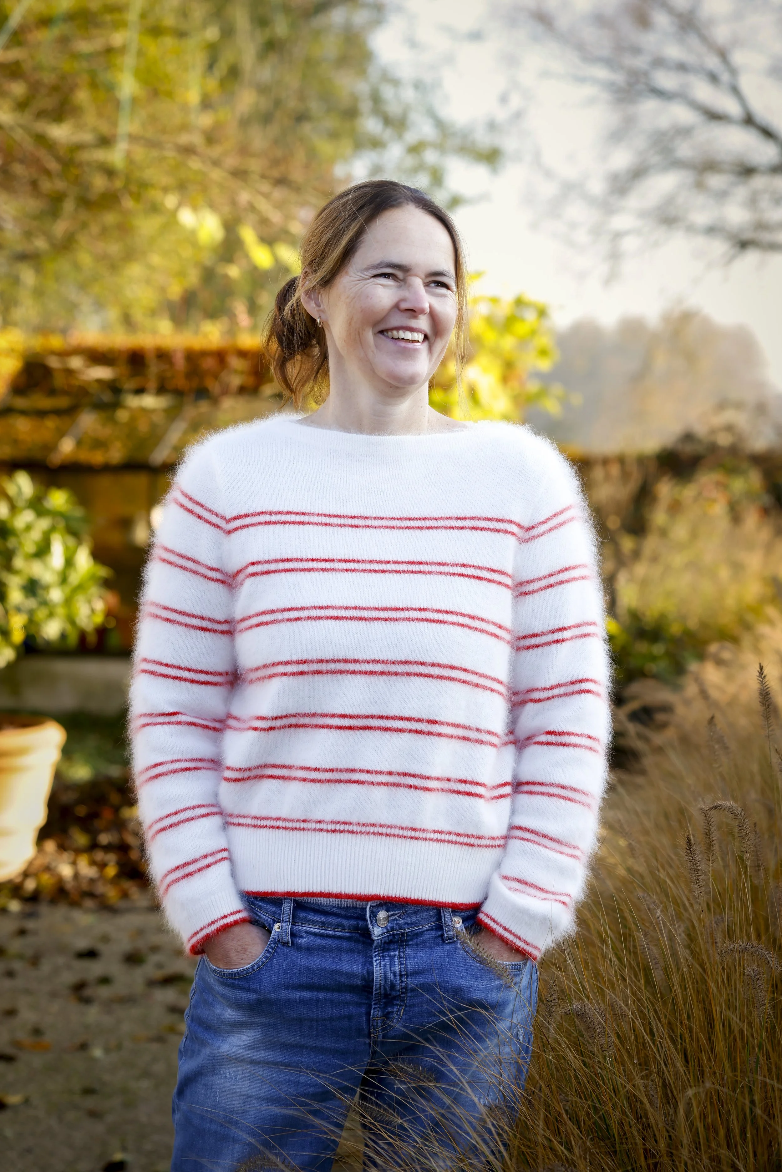 Vrouw staat glimlachend in een tuin met herfstbladeren.