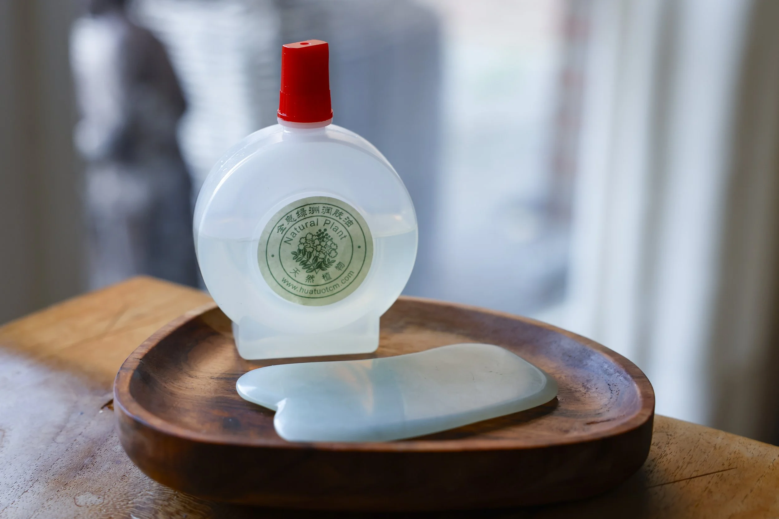 A bottle of clear liquid with a green label and red cap sits on a wooden tray, next to a jade roller. The label has Chinese characters and the words 'Natural Plant' and a website.