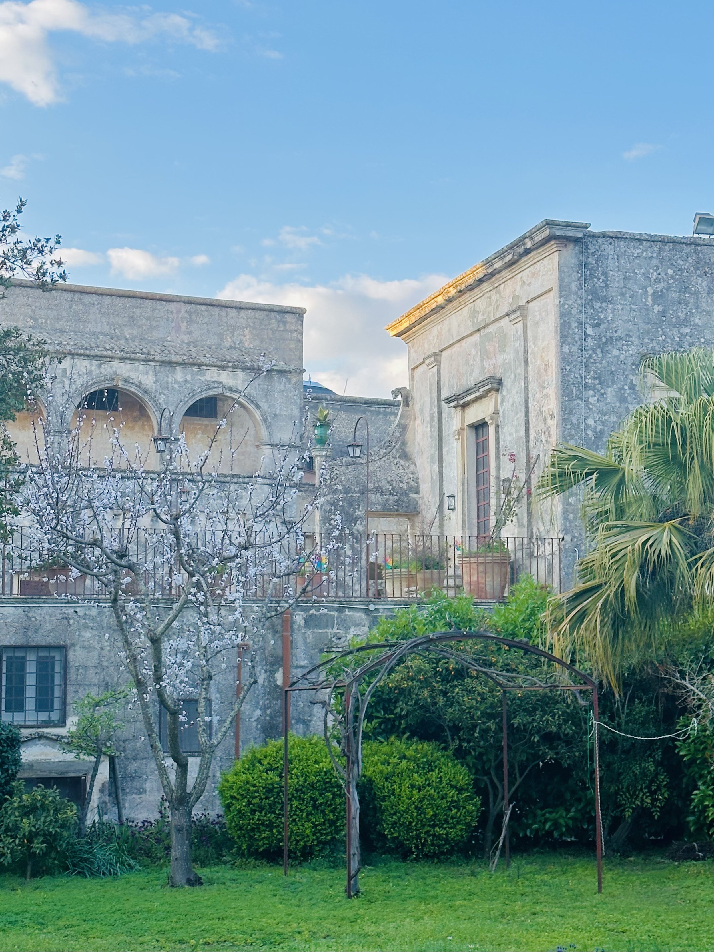 View of the historic facade and arched gallery of Palazzo Bacile in Spongano, Puglia, as seen from the historical citrus garden, surrounded by lush trees and greenery under a clear blue sky