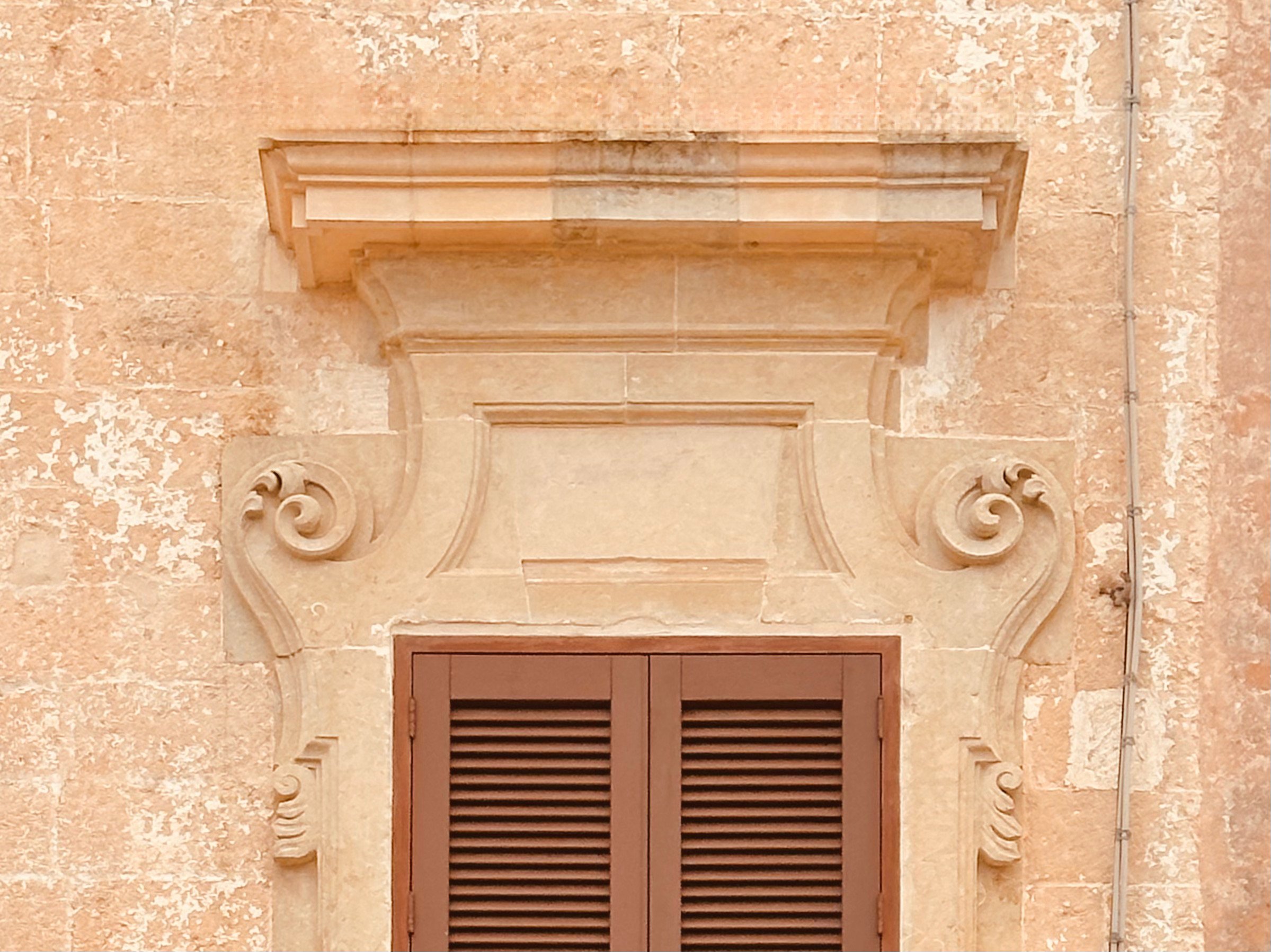Ornate stone window frame on the historic facade of Palazzo Bacile in Spongano, Puglia, with closed wooden shutters reflecting traditional southern Italian architecture