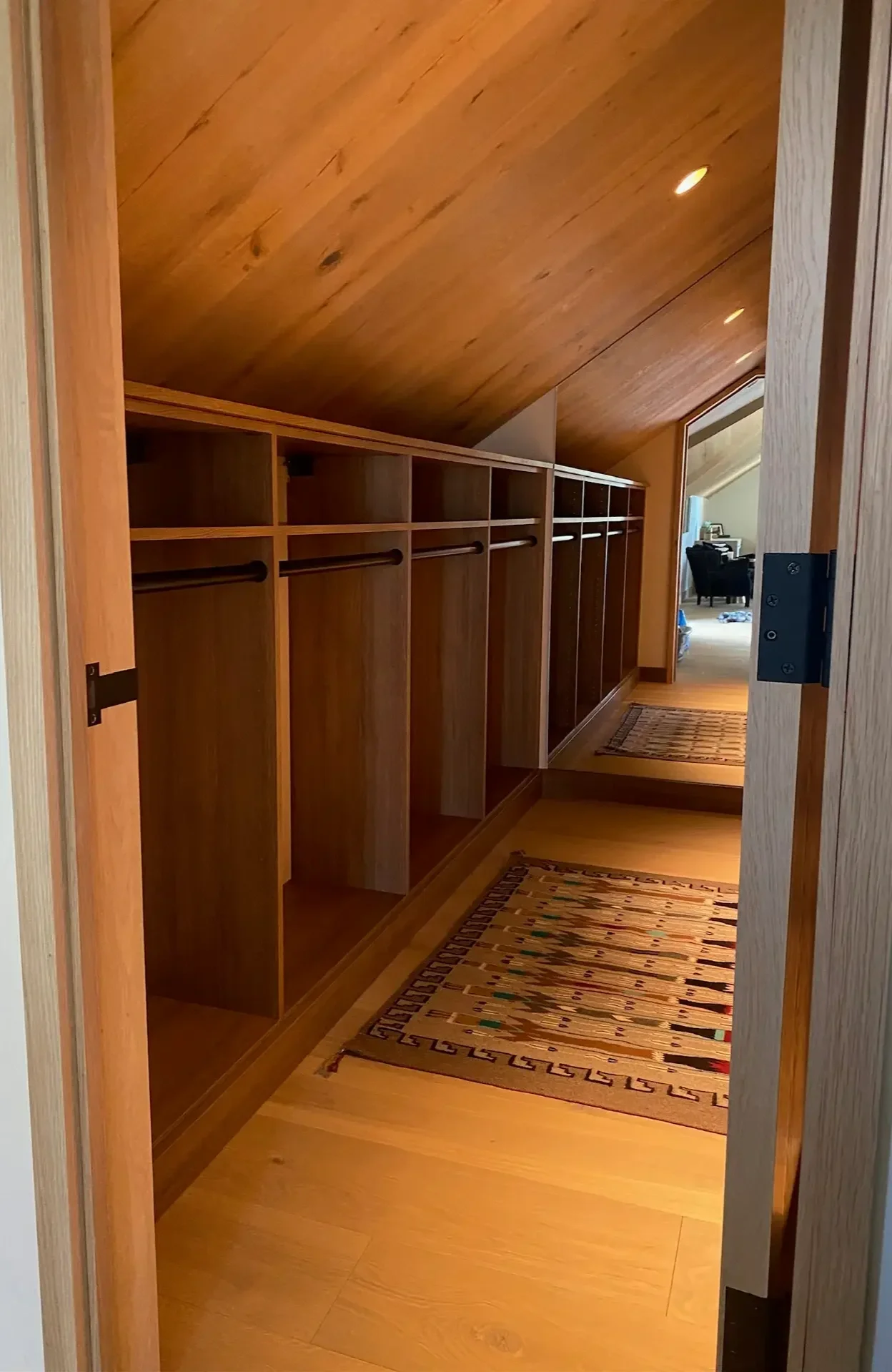 A walk-in closet with wooden shelving and an angled wooden ceiling. The floor is wood, and there is a colorful patterned rug on it. In the background, a living room with a gray couch and armchair, a small table, and stockings hanging on the wall are visible.