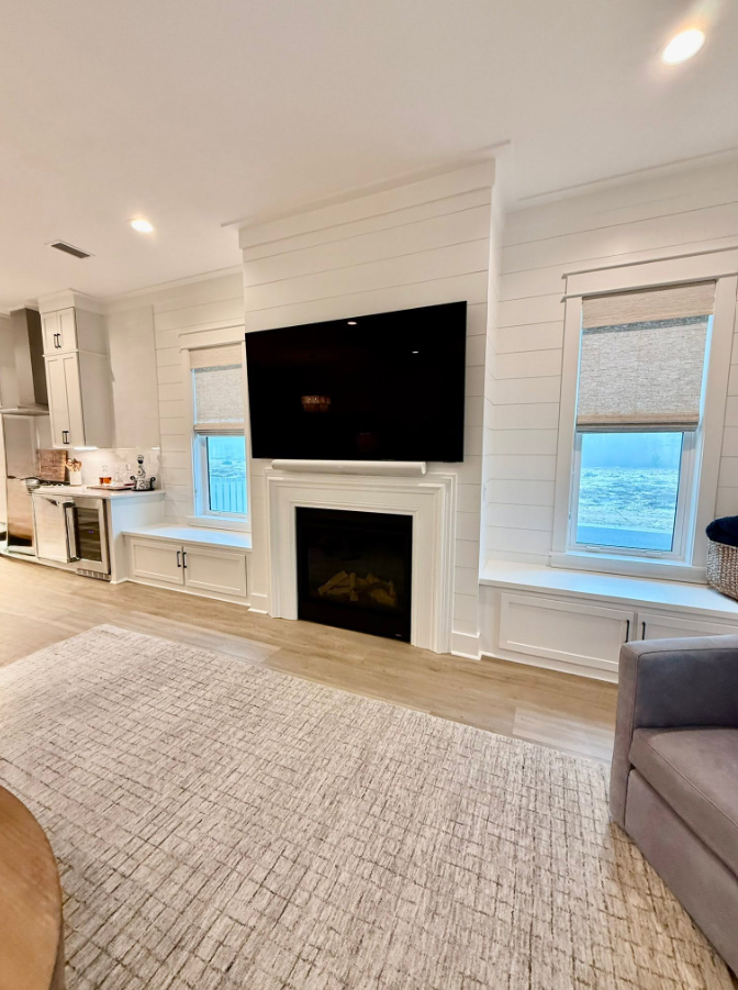 Living room with white walls, a fireplace with a black screen above, two windows with shades, a beige rug, and a grey armchair.