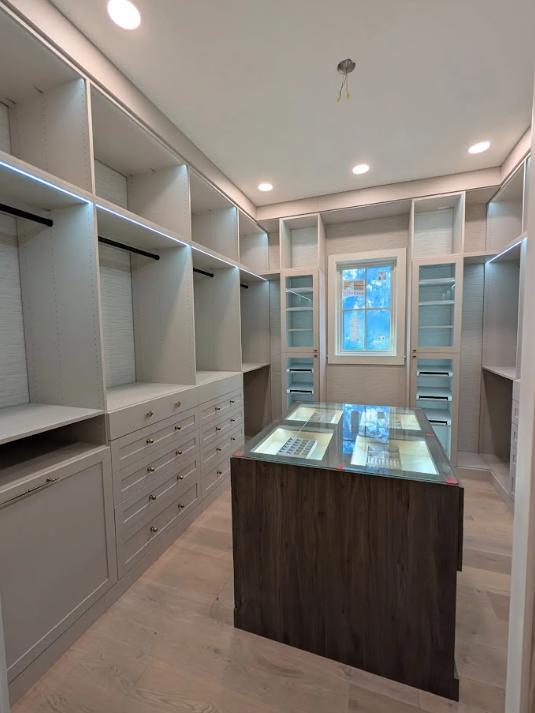 Empty walk-in closet with built-in shelves, drawers, and a central island with a glass top, illuminated by ceiling lights and a window at the back.
