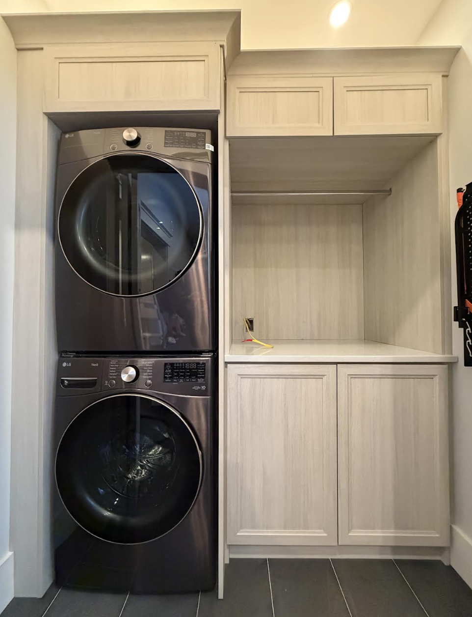 Stacked front-loading washer and dryer in a laundry area with light-colored cabinets above and to the right, with a black tile floor.