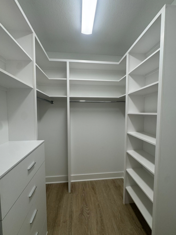 Empty white walk-in closet with shelves and drawers, wood floor, ceiling light.