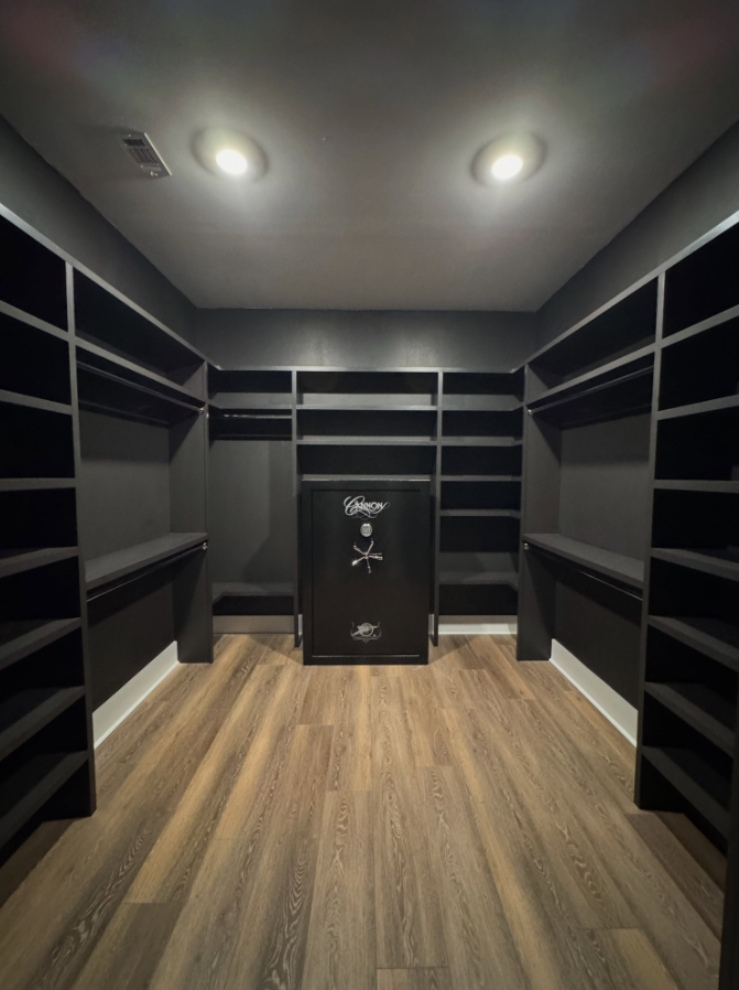 A walk-in closet with dark wooden shelves and a small black safe in the center, illuminated by two ceiling lights.