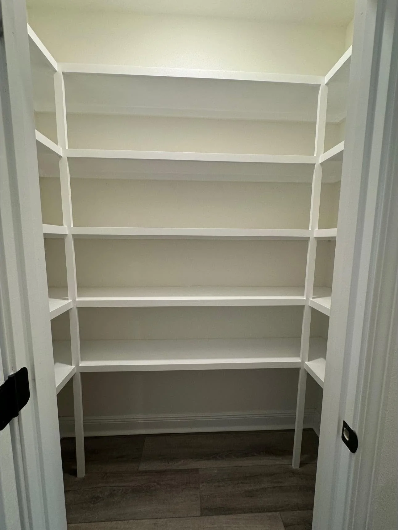Empty white built-in pantry with multiple shelves, open double doors, and dark wood laminate flooring.