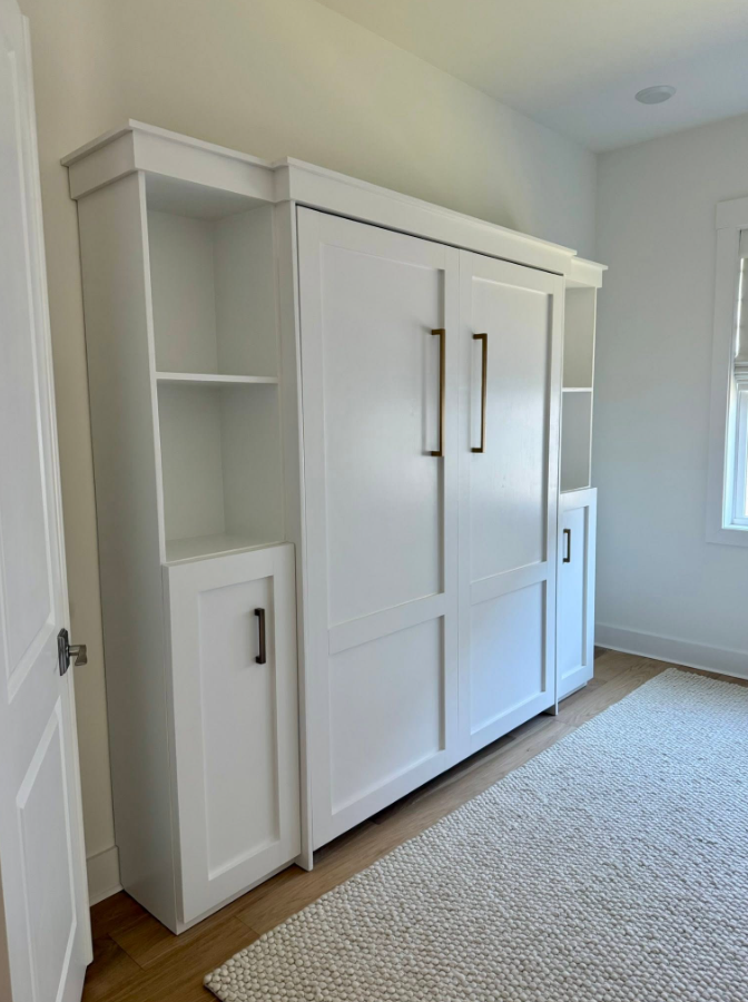 White built-in storage cabinet with doors, shelves, and brass handles in a bright room with hardwood flooring and a window.