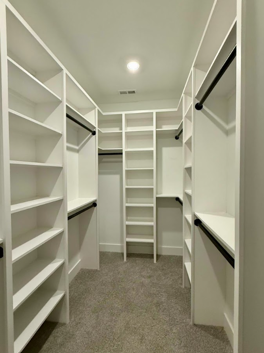 Empty walk-in closet with white shelves, hanging rods, and gray carpeted floor.