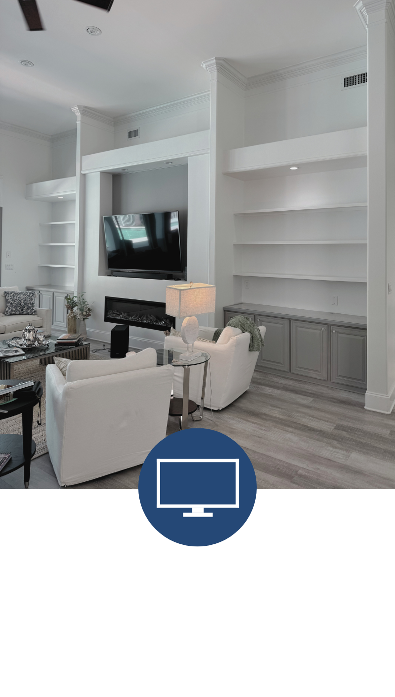 Modern living room with white walls, a mounted flat-screen TV above a fireplace, white armchairs, a side table with a lamp, built-in shelves, and gray cabinetry.