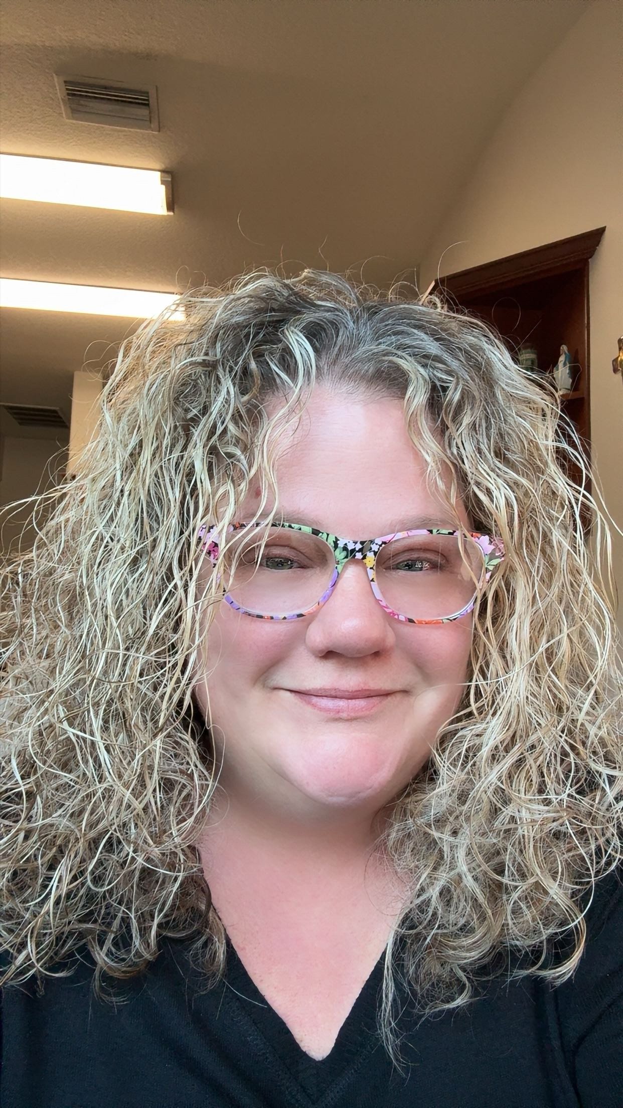 A woman with curly blonde hair and colorful patterned glasses smiling indoors.
