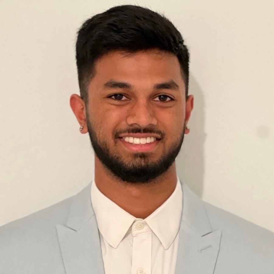 A young man with dark hair and a beard, wearing a light gray suit and white shirt, smiling in front of a plain light background.