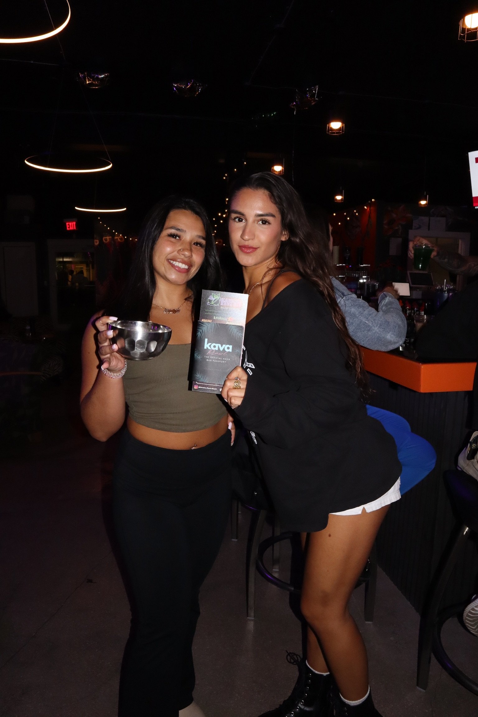 Two women holding a brochure and a glass in a bar, smiling at the camera.