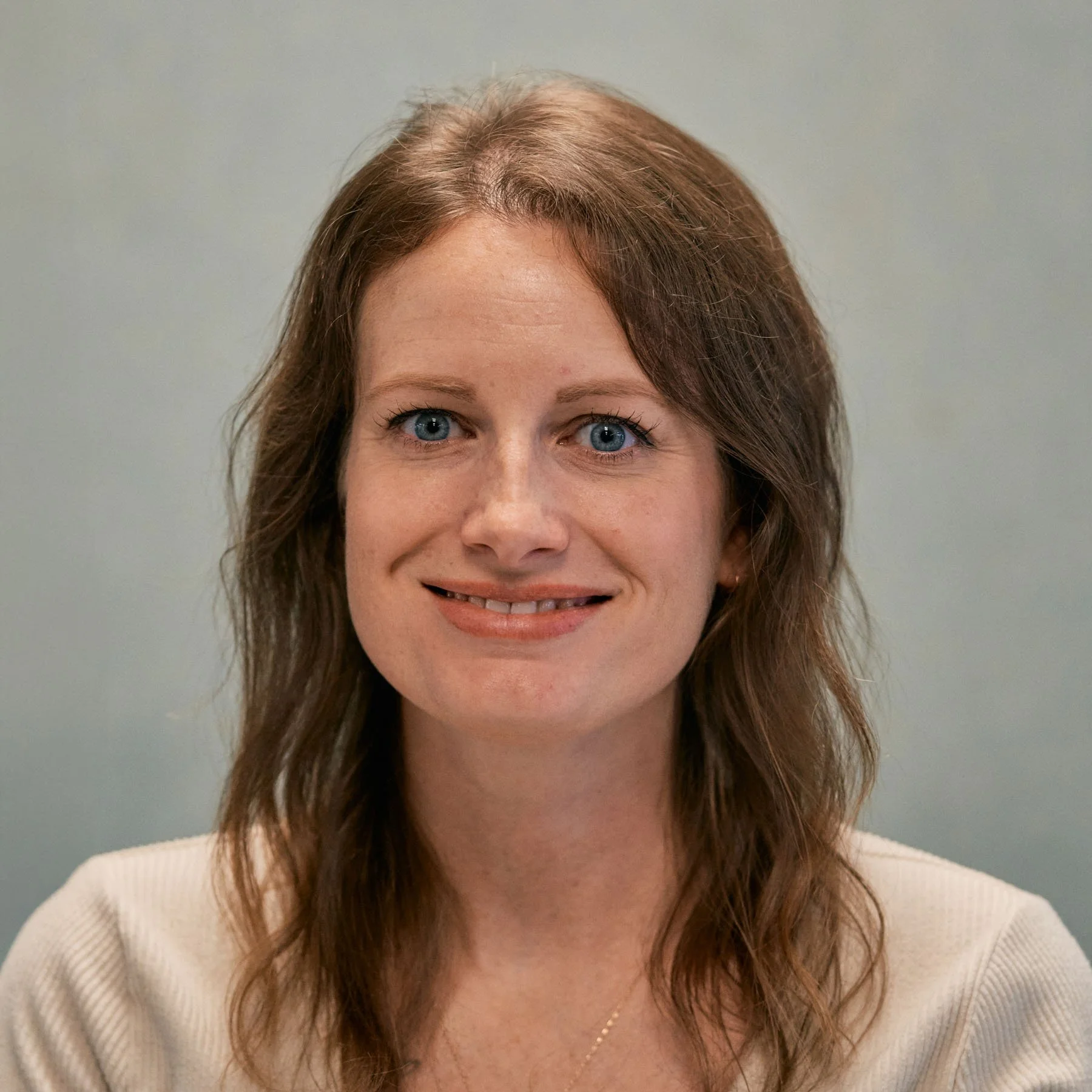A woman with long brown hair smiling at the camera against a beige background, wearing an olive green jacket over a white shirt with black stripes.