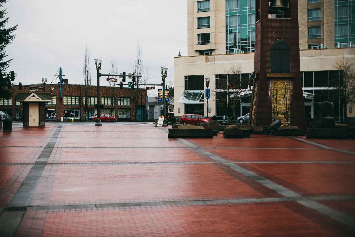 Empty plaza with brick paving, a few parked cars, and surrounding buildings including a multi-story building with glass windows, trees, streetlights, and traffic signals