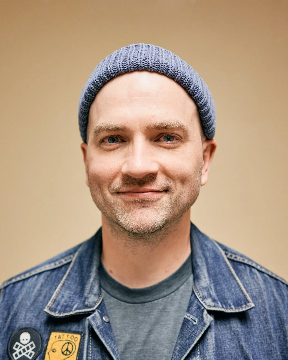 A young man with blue eyes, wearing a blue knit beanie and a denim jacket, smiling at the camera against a beige background.