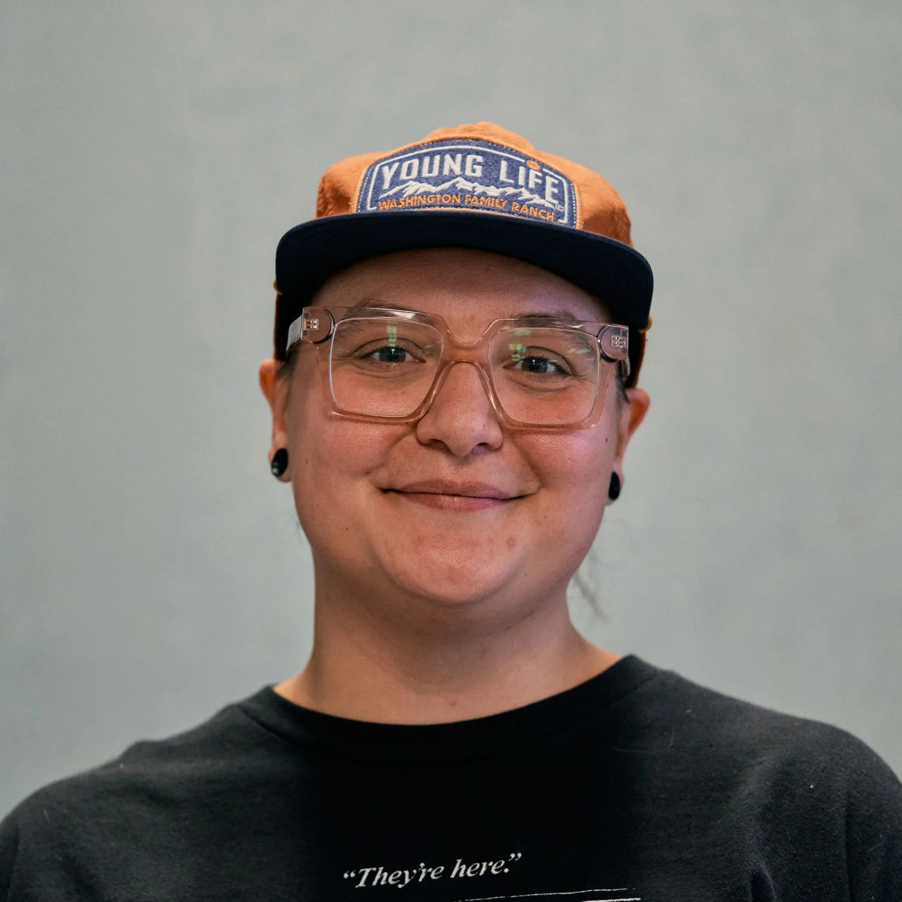 A woman with wavy, light brown hair and glasses, smiling indoors against a plain beige wall.