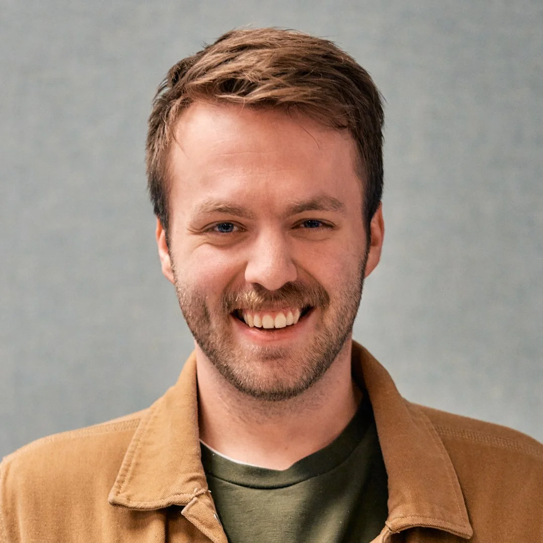 Close-up of a smiling young man with brown hair, blue eyes, and a light beard, wearing a beige shirt, against a neutral background.