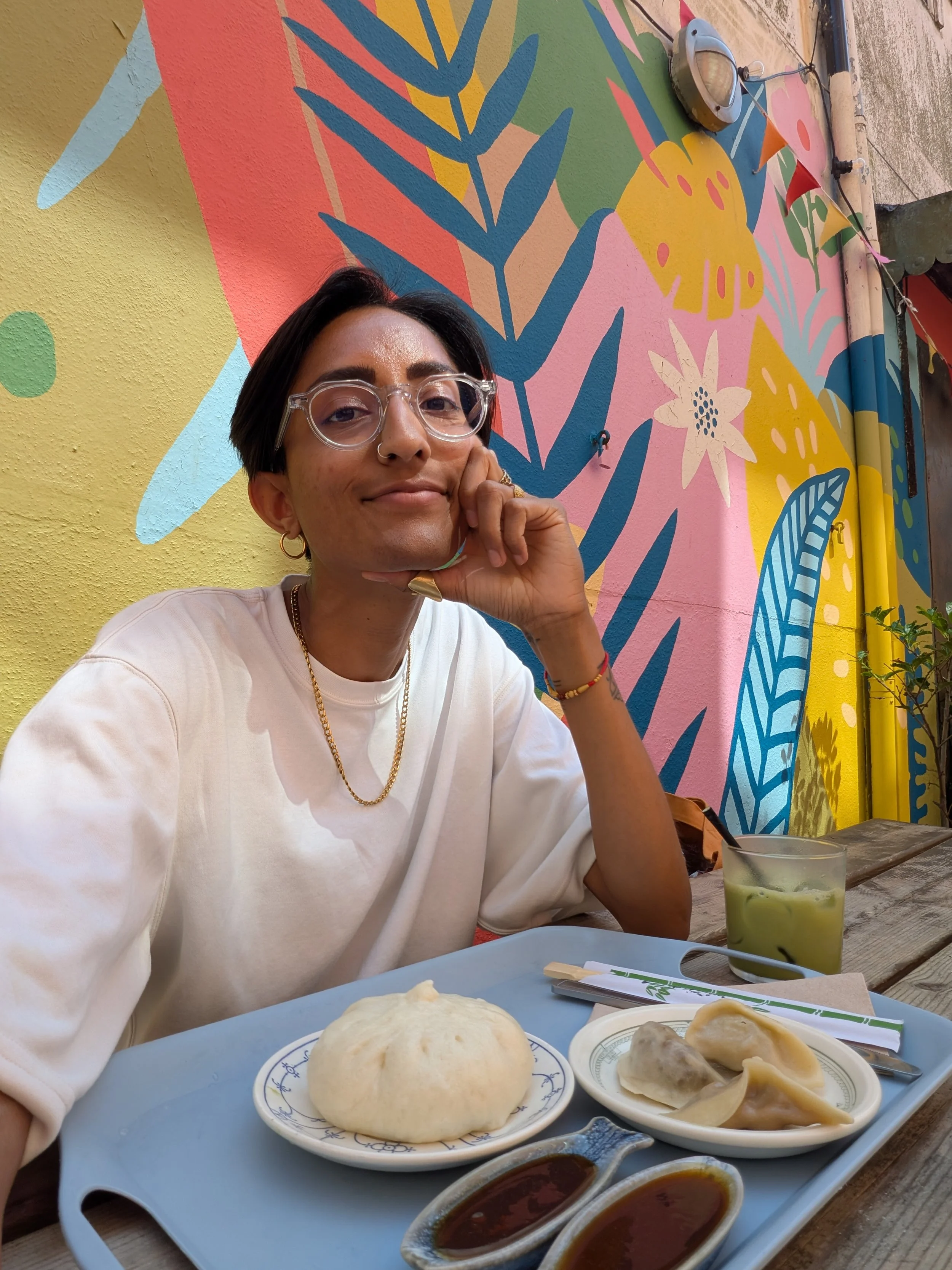 A picture of Henna wearing a white t-shirt and glasses. With food on the table in front of them and their left hand on their cheek.