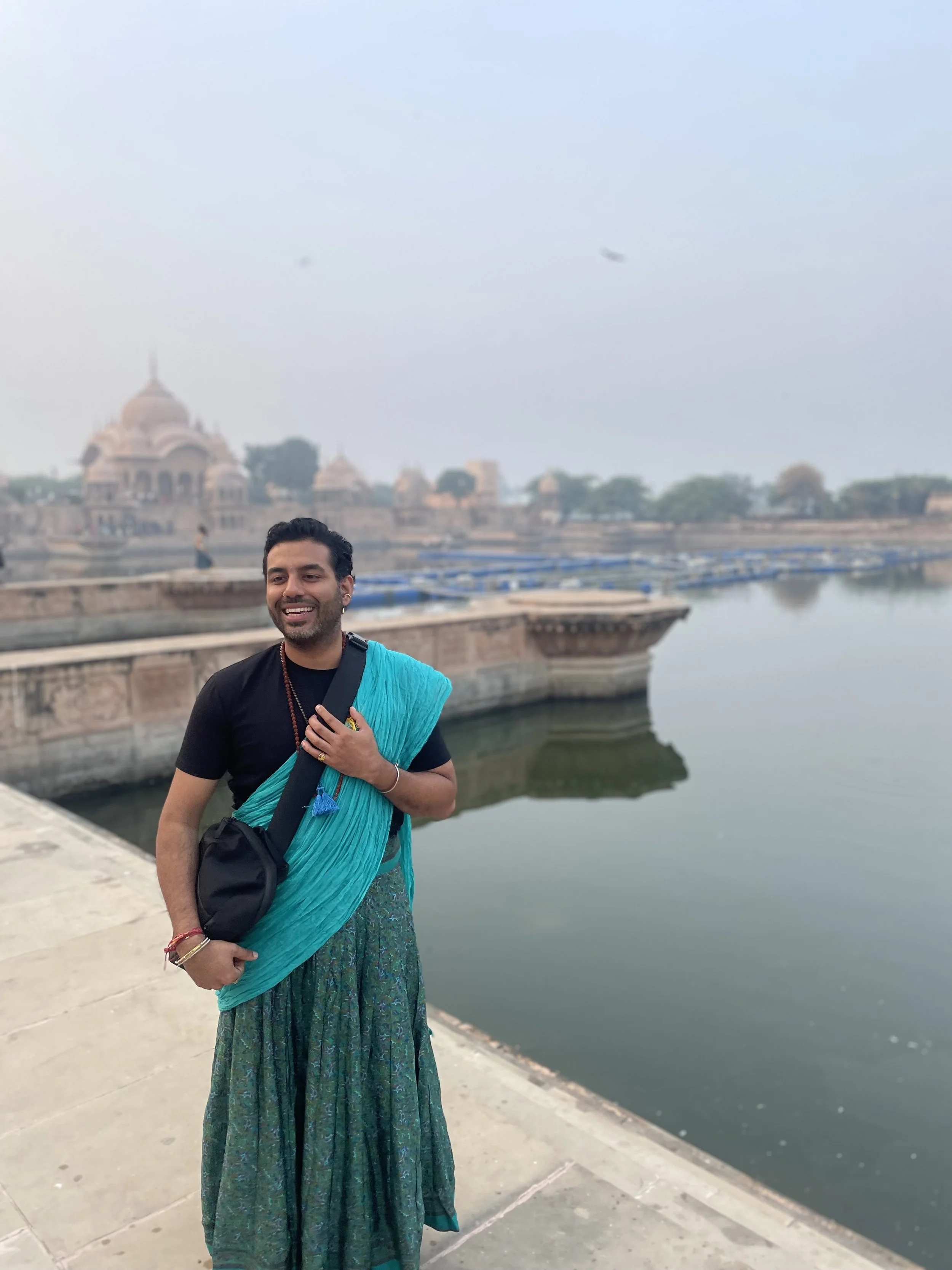 An image of Shay wearing a peacock coloured gopi dress and a black t-shirt in front of a palace and lake in India, Vrindavan