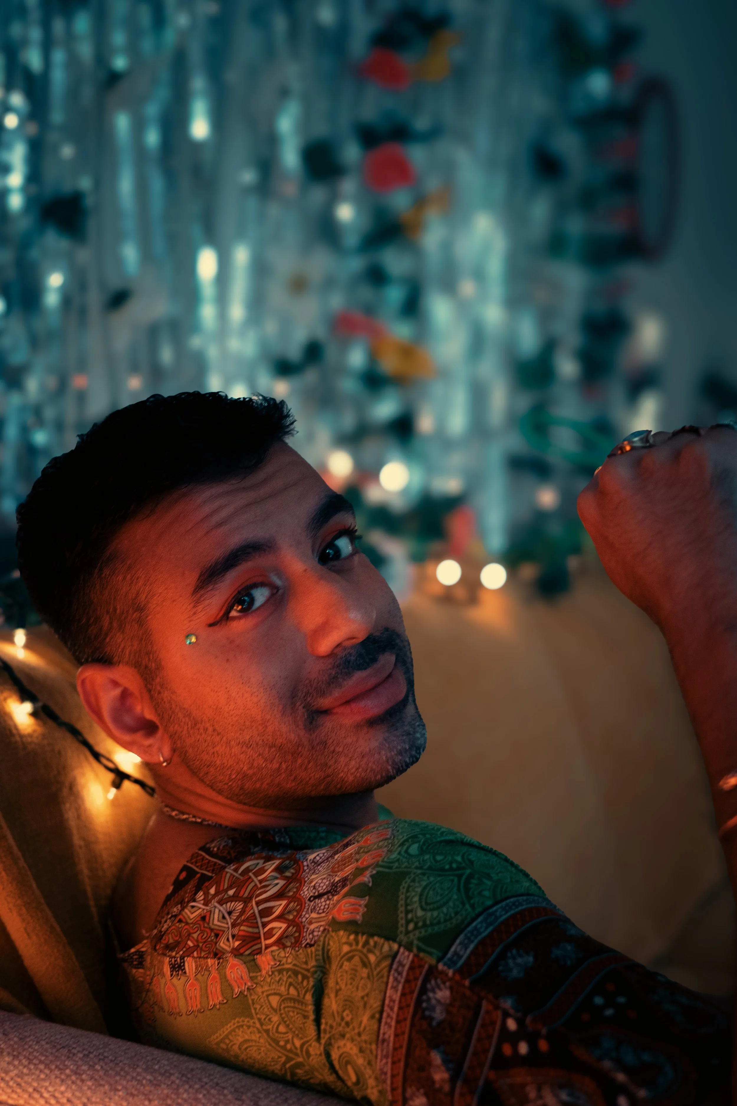 A profile image of Shay sitting on a couch and looking back over their shoulder at the camera, with short black hair and orange eye shadow