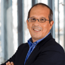 Ein Mann in formeller Kleidung, trägt Brille und lächelt, vor einem blauen und grauen Hintergrund.