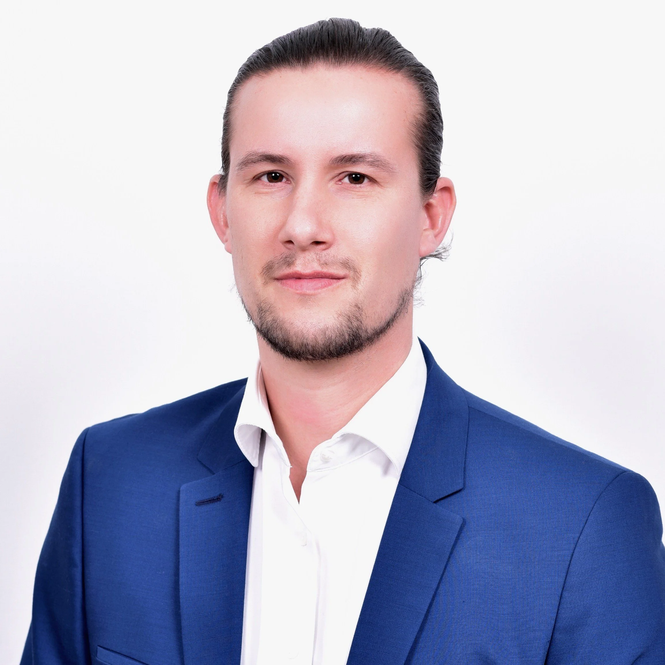 Porträt eines Mannes mit dunklem Haar, heller Haut, blauer Anzug und weißem Hemd vor weißem Hintergrund.