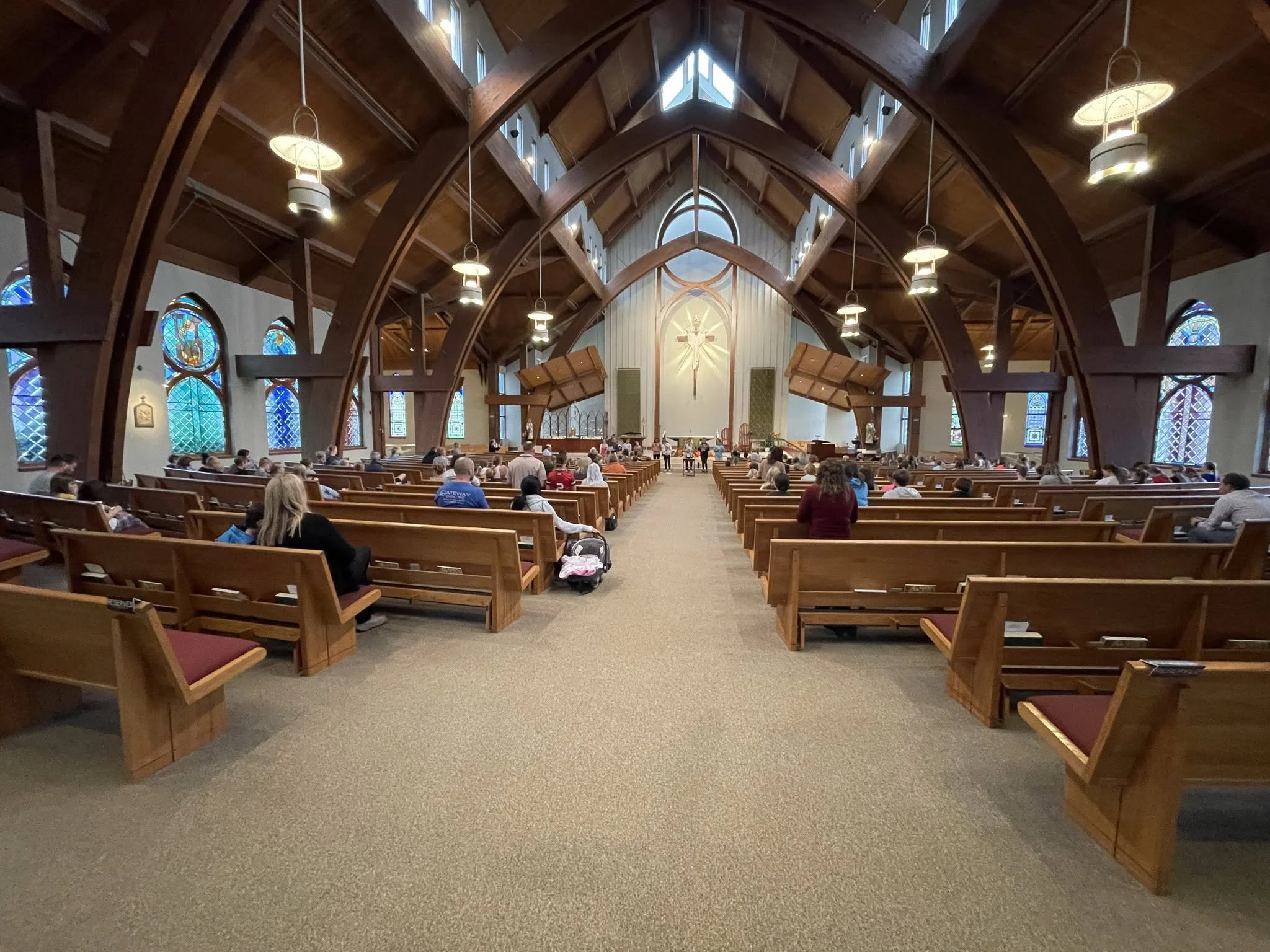 Interior view of a church with wooden beams, stained glass windows, and rows of pews filled with people attending a service.