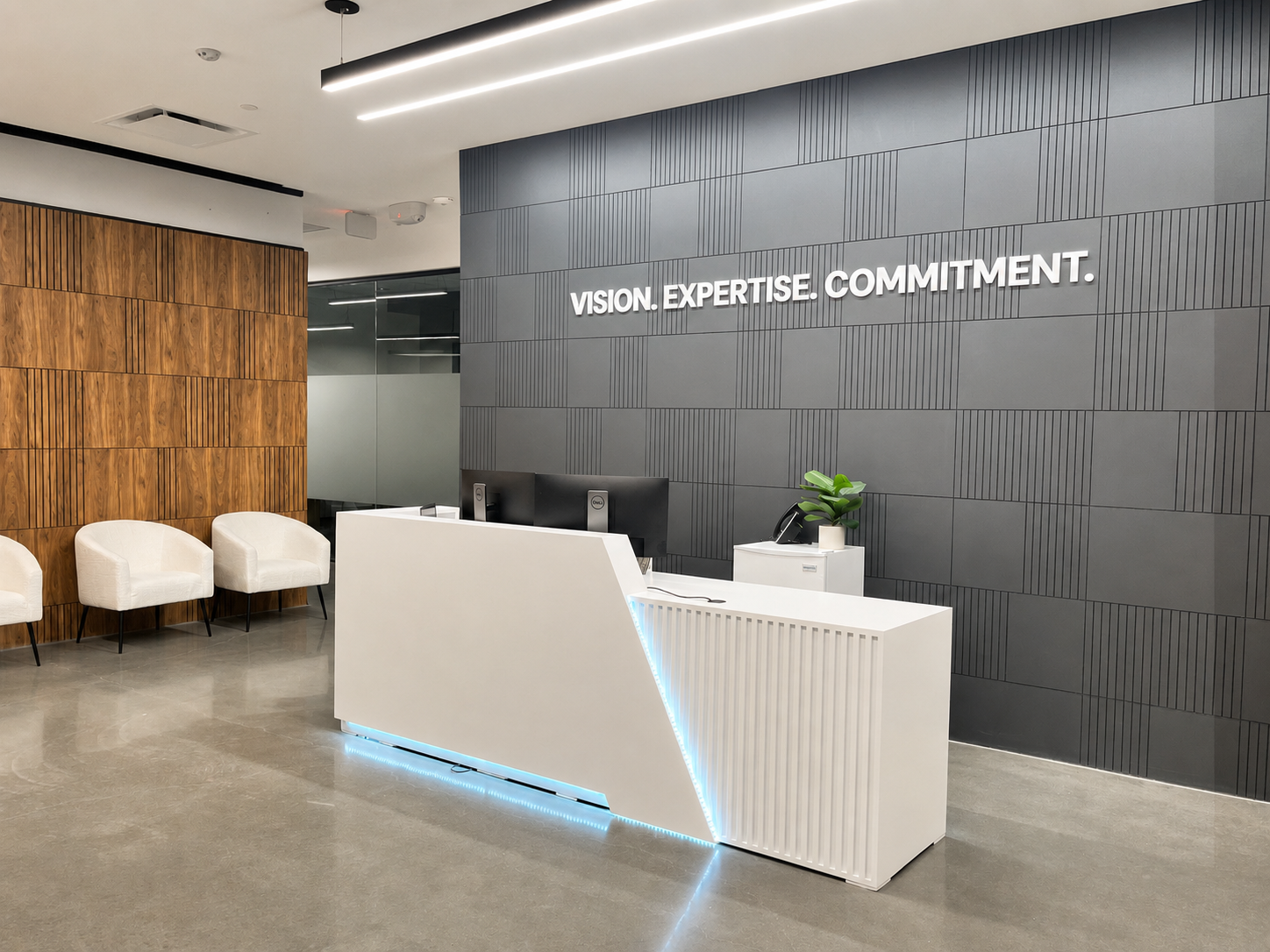Modern office reception area with a white front desk, a black computer monitor, a potted plant, and a gray wall with the words 'Vision. Expertise. Commitment.' in white letters. There are four white chairs against a wooden wall to the left.